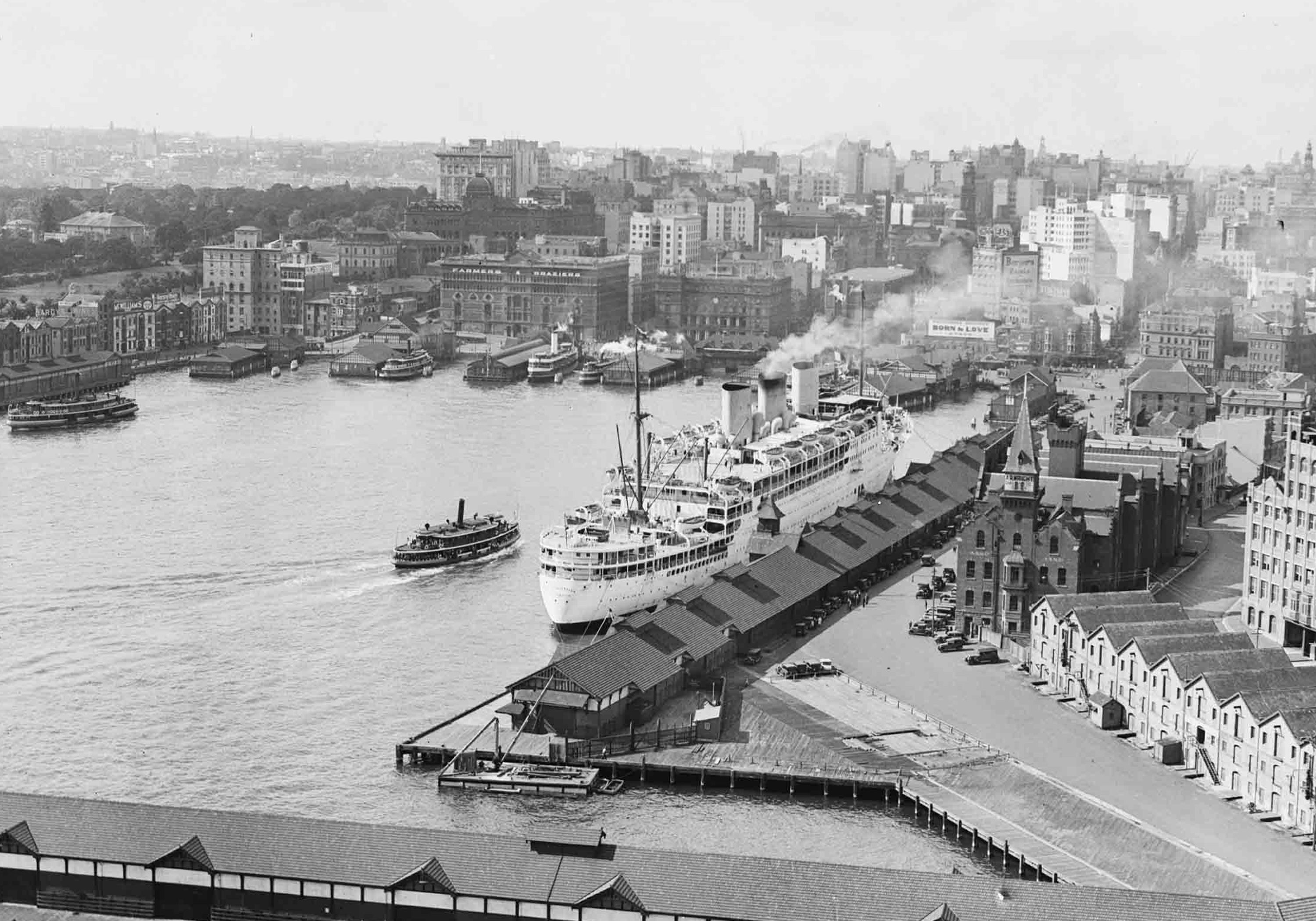 A black and white photograph taken from a high elevation down on a harbour, with a large white cruise ship at the dock closest to the camera an many other smaller boats in the water and at other docks. The background is an old city.