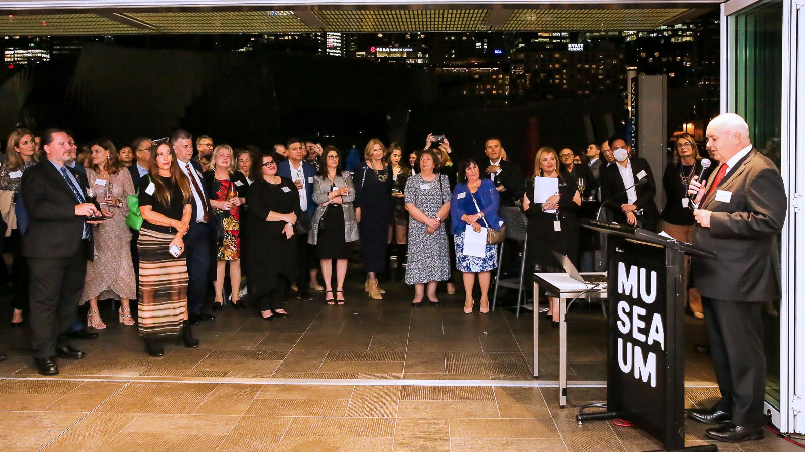 Photo of a crowd of well dressed people watching a presenter who is a man wearing a suit and standing behind a lecturn at the right side of the photo.