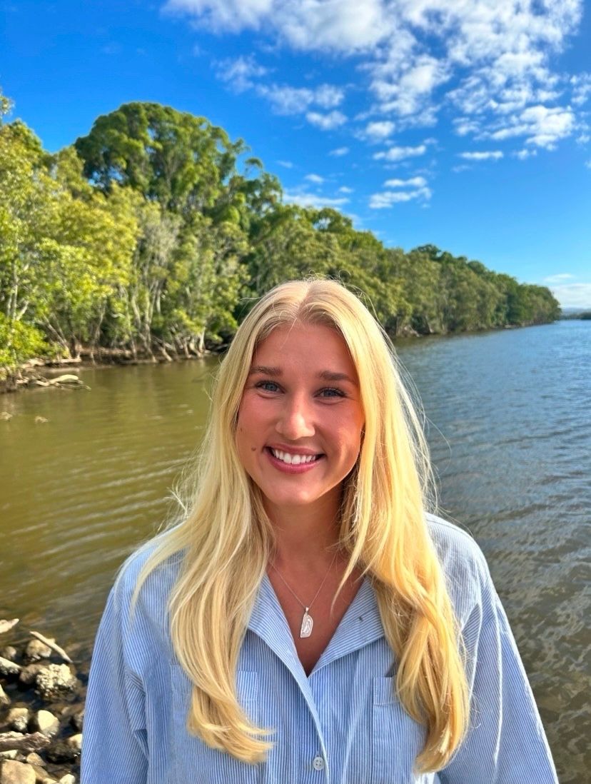 Woman with long blonde hair smiling in a blue button shirt in front of a creek with blue skies