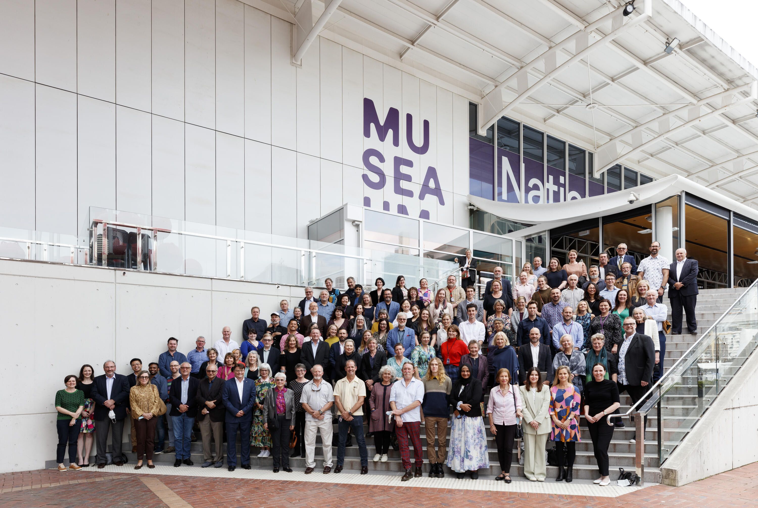 large group of people standing on wide steps in front of a white building with the museum logo on it.