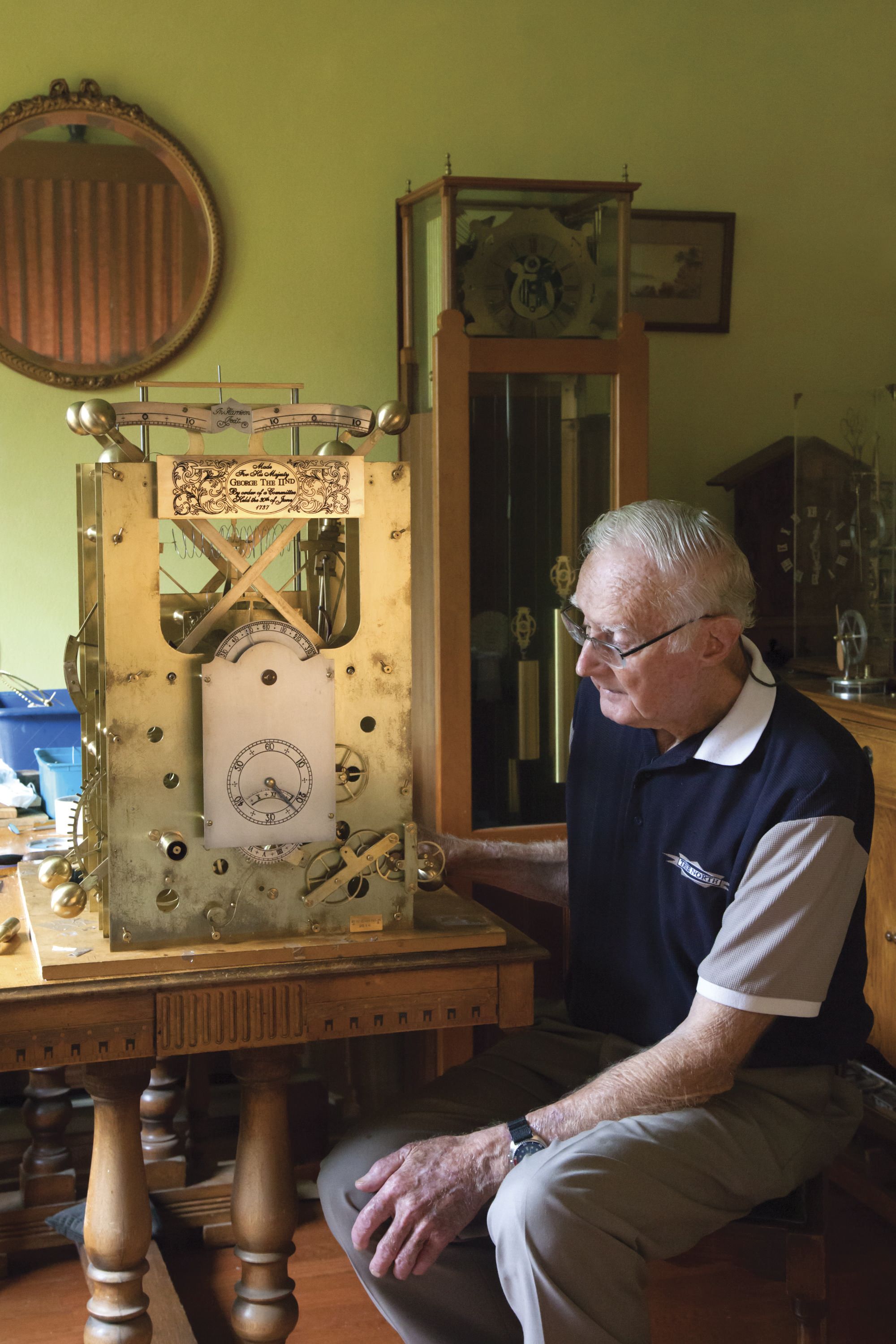Photograph showing an older man sitting next to a large, elaborate clock. 
