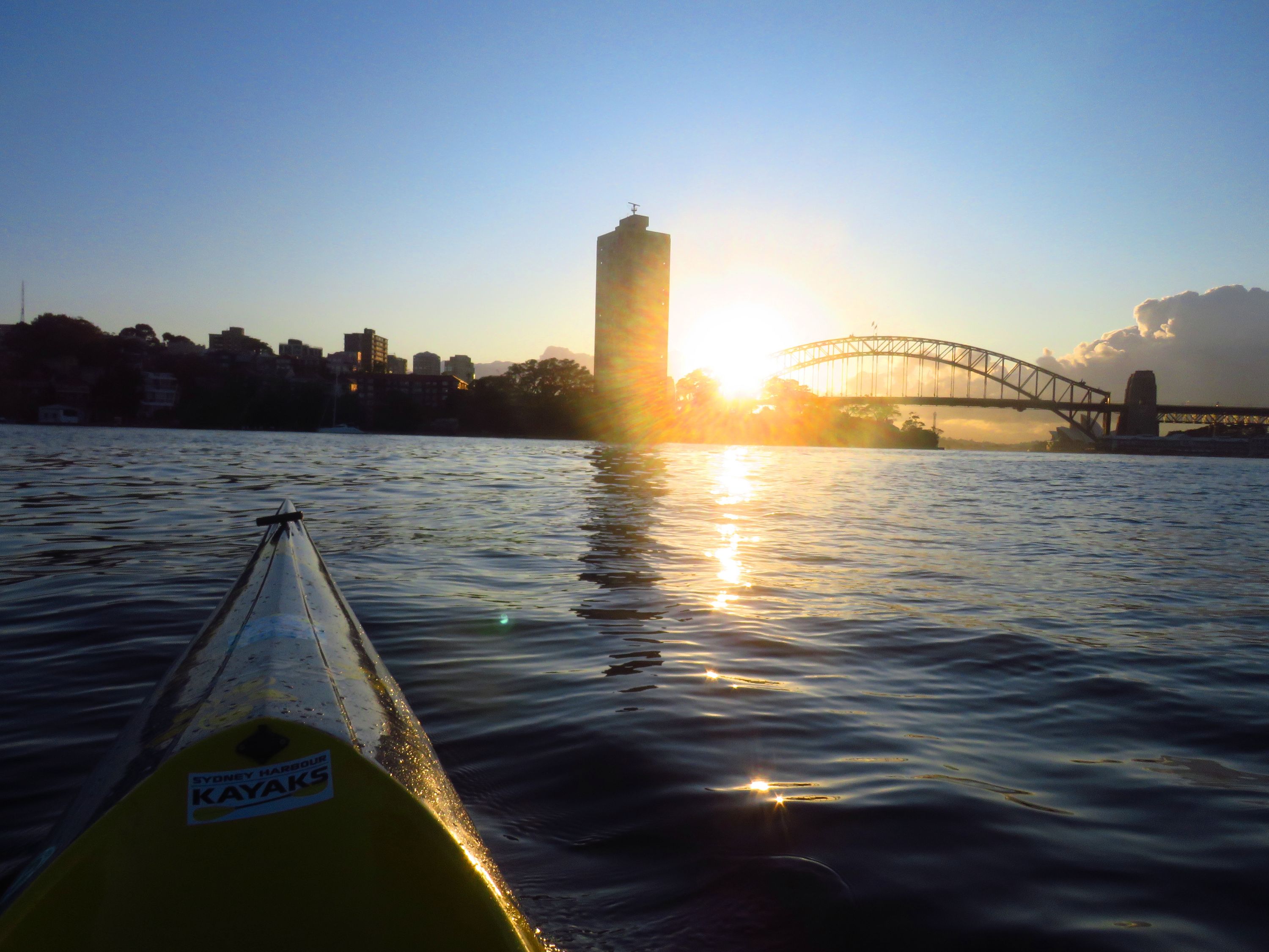 Photograph taken from on a kayak, looking towards the sun rising behind the Sydney harbour bridge and a skyscraper building. 