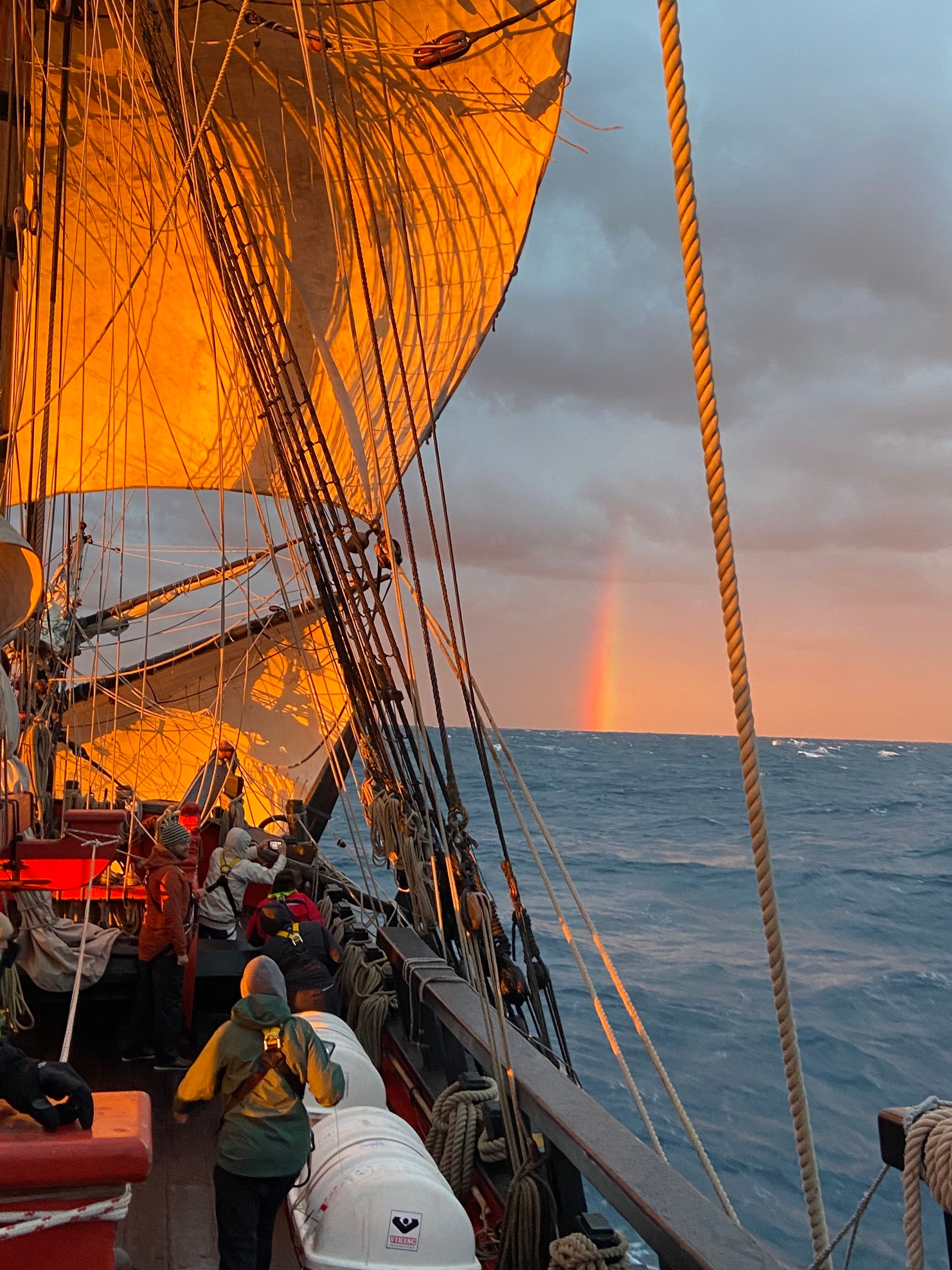 Photo from the deck of a tall ship sailing, the sails in golden orange light, part of a rainbow in the cloudy sky above the blue sea.