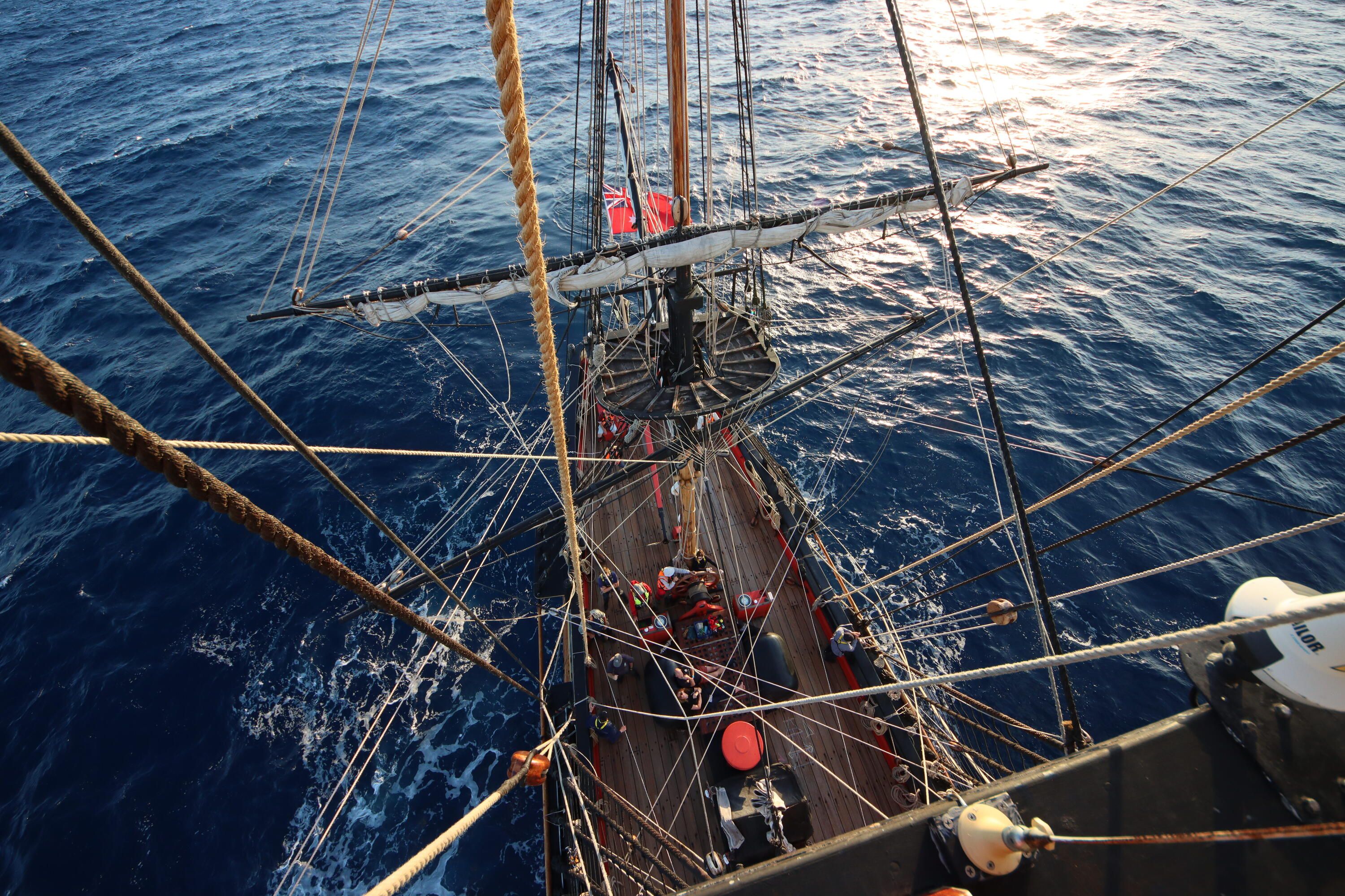 A photo taken from up the top of a mast looking down at a tall ship moving through the water. 