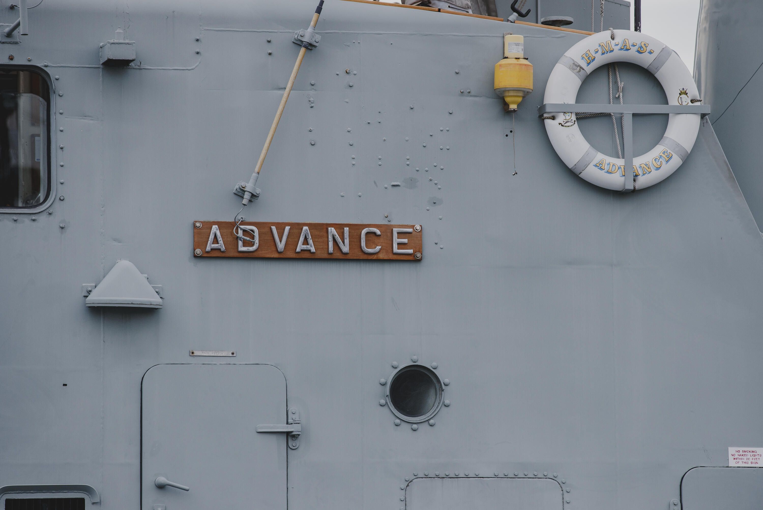 Close up photo showing the side of a Navy ship, with a wooden plaque saying "ADVANCE".