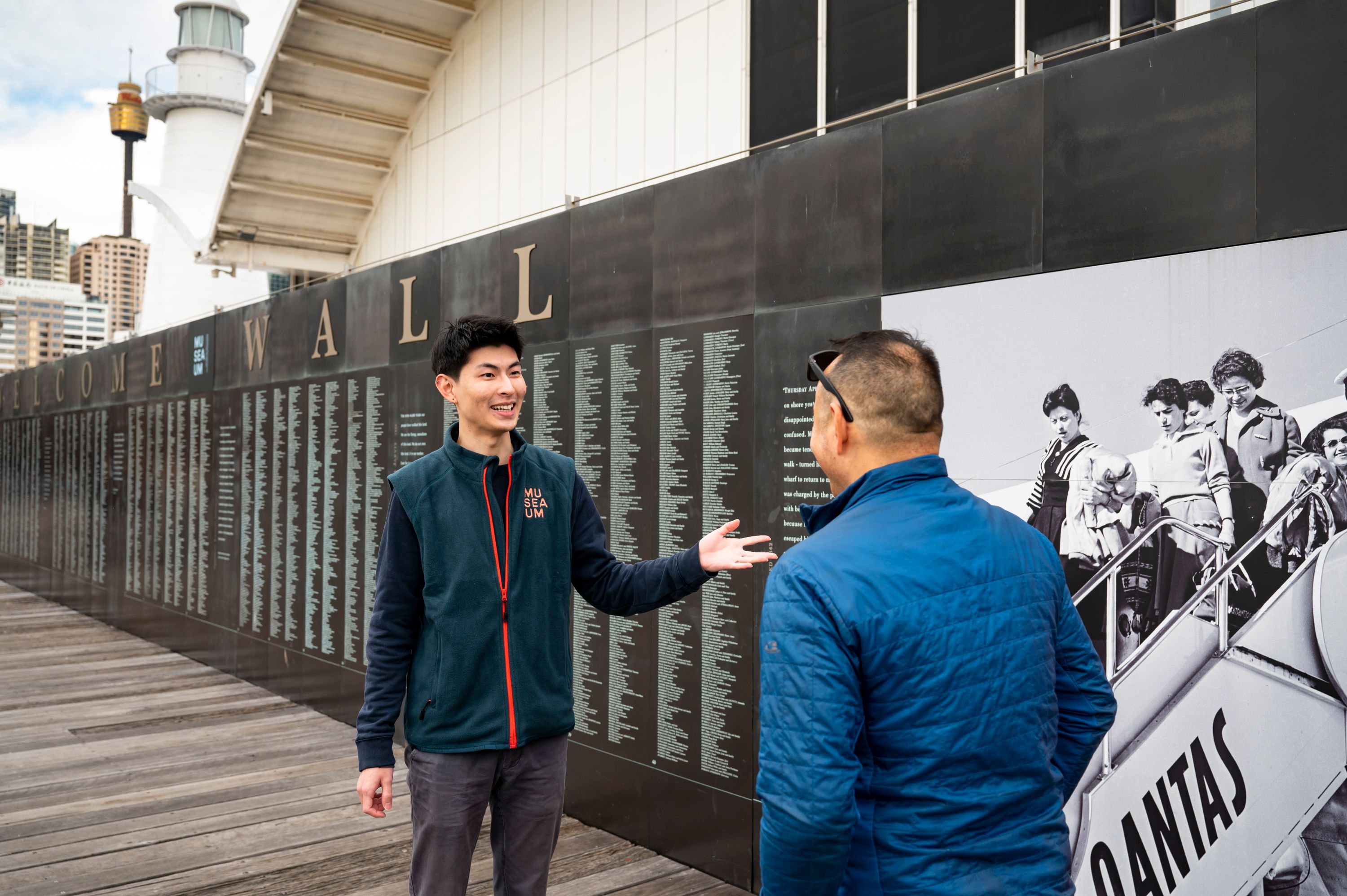 Male volunteer in a uniform showing a visitor names engraved on the national monument to migration