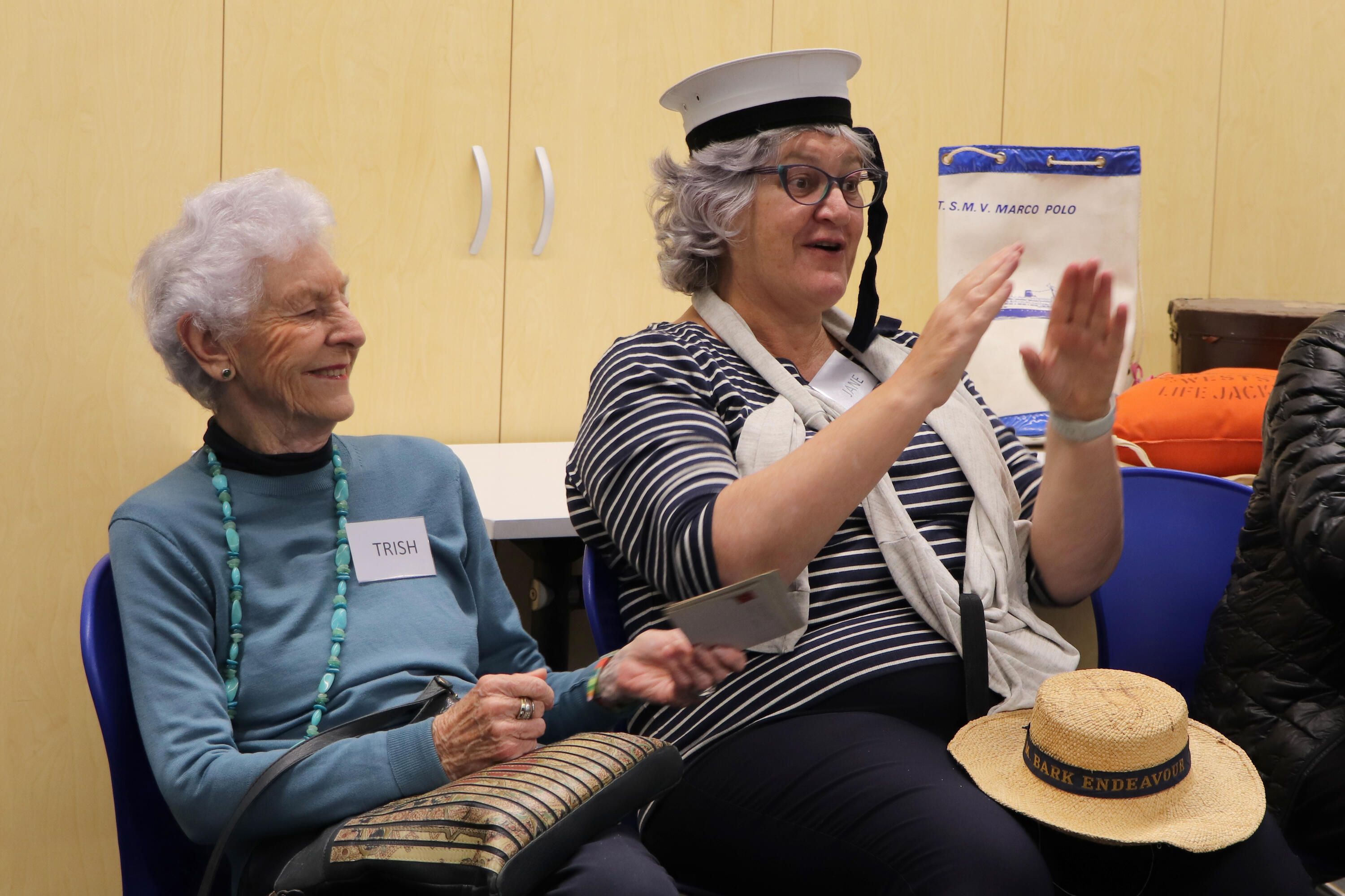A lady with short white hair and a blue jumber sitting next to another lady with a stripped top and sailor's hat.