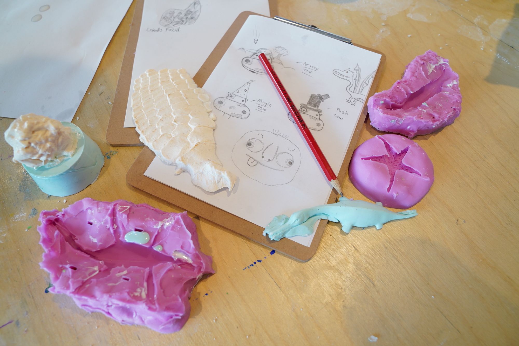 A table with a clipboard with paper containing pencil drawings, surrounded by pink silicone moulds 