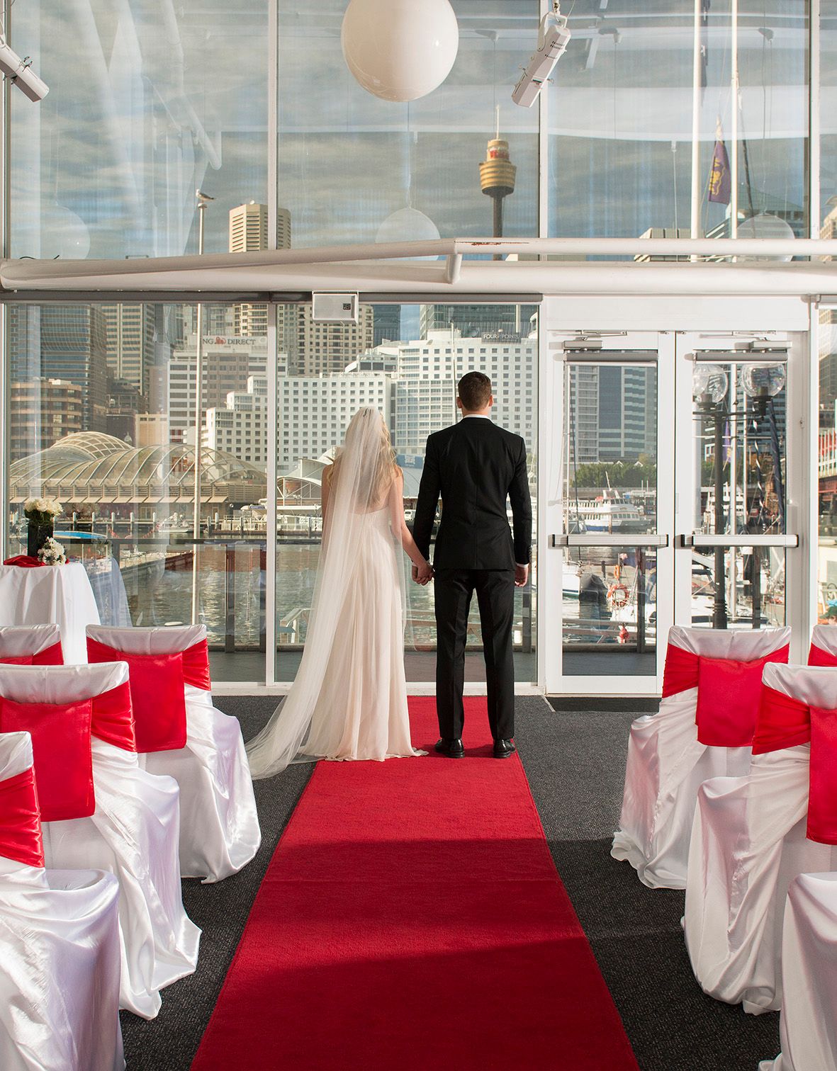 Photo showing the backs of a bride and groom standing at the end of a red carpet, in front of a window with city views.