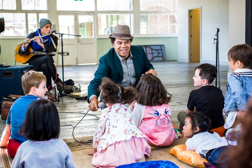 Photo of a man wearing a hat, surrounded by a group of children and a guitar player in the background
