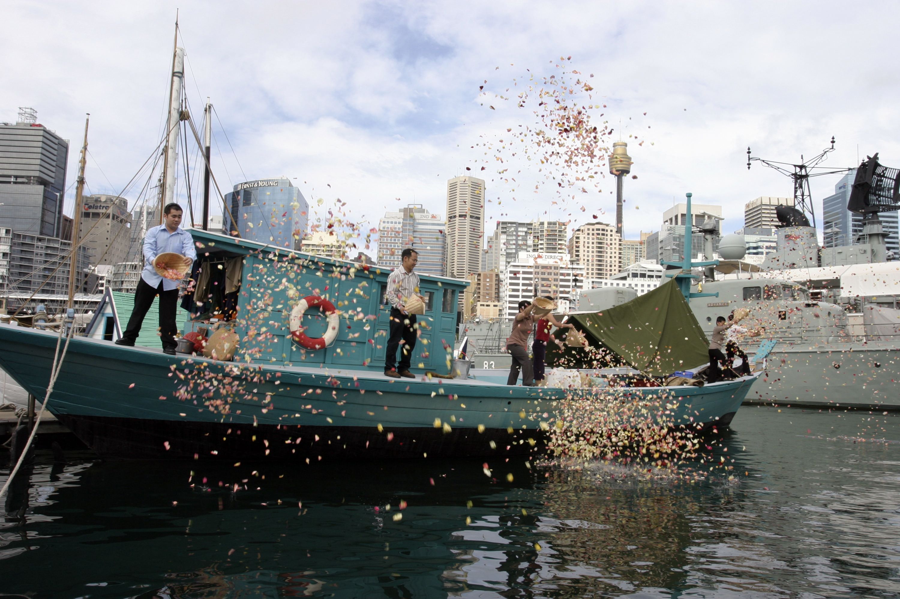Blue Fishing Boat. Standing on board are the Lu family members who came to Australia from Vietnam as refugees on board the boat. They are scattering flower petals into the harbour in celebration.
