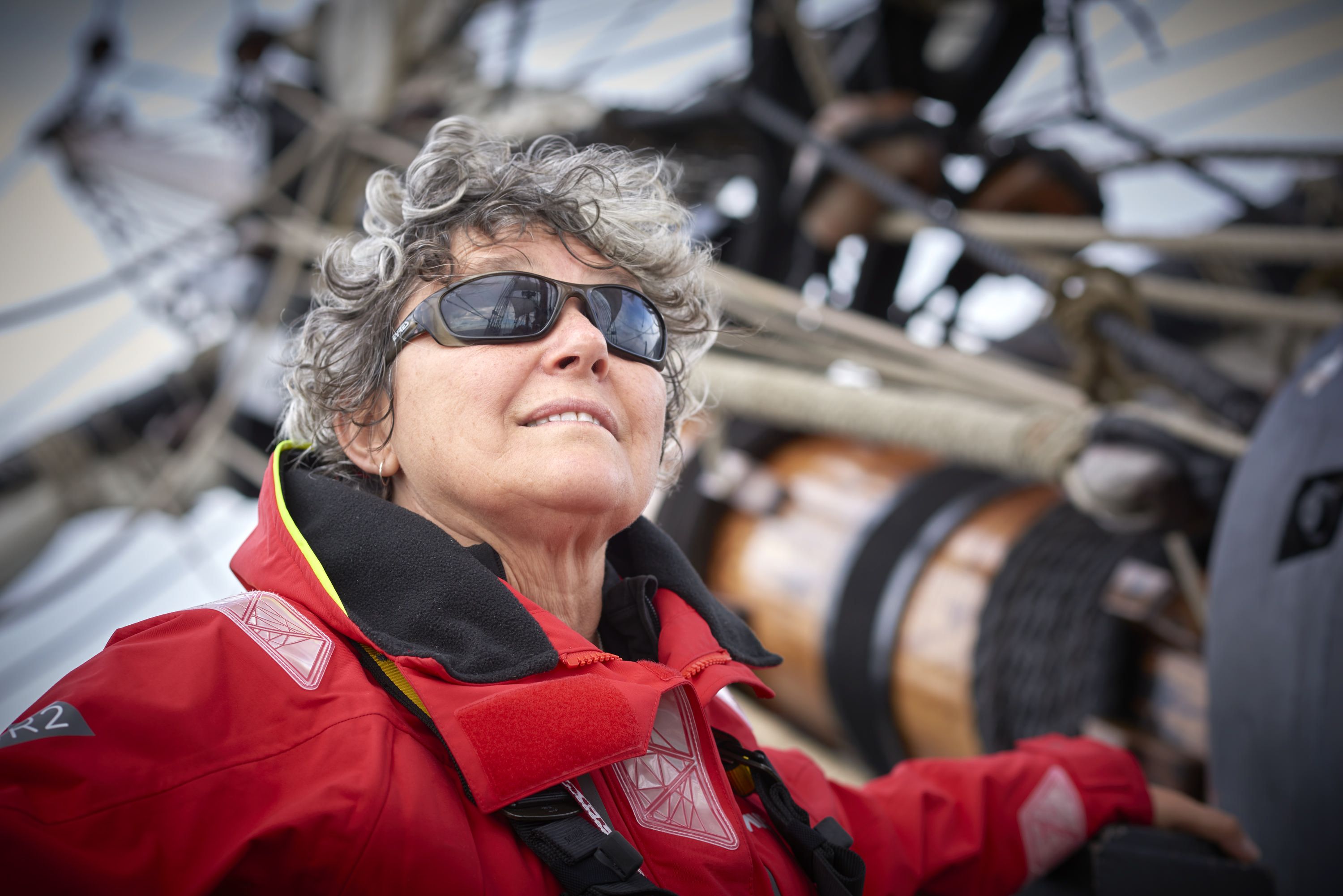 Above deck, woman wearing sunglasses looking out to sea with mast in background. 