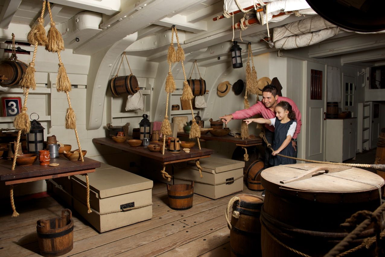 Photo showing below decks of a historical ship, with wooden tables, trunks and buckets. A man and girl are looking and pointing. 