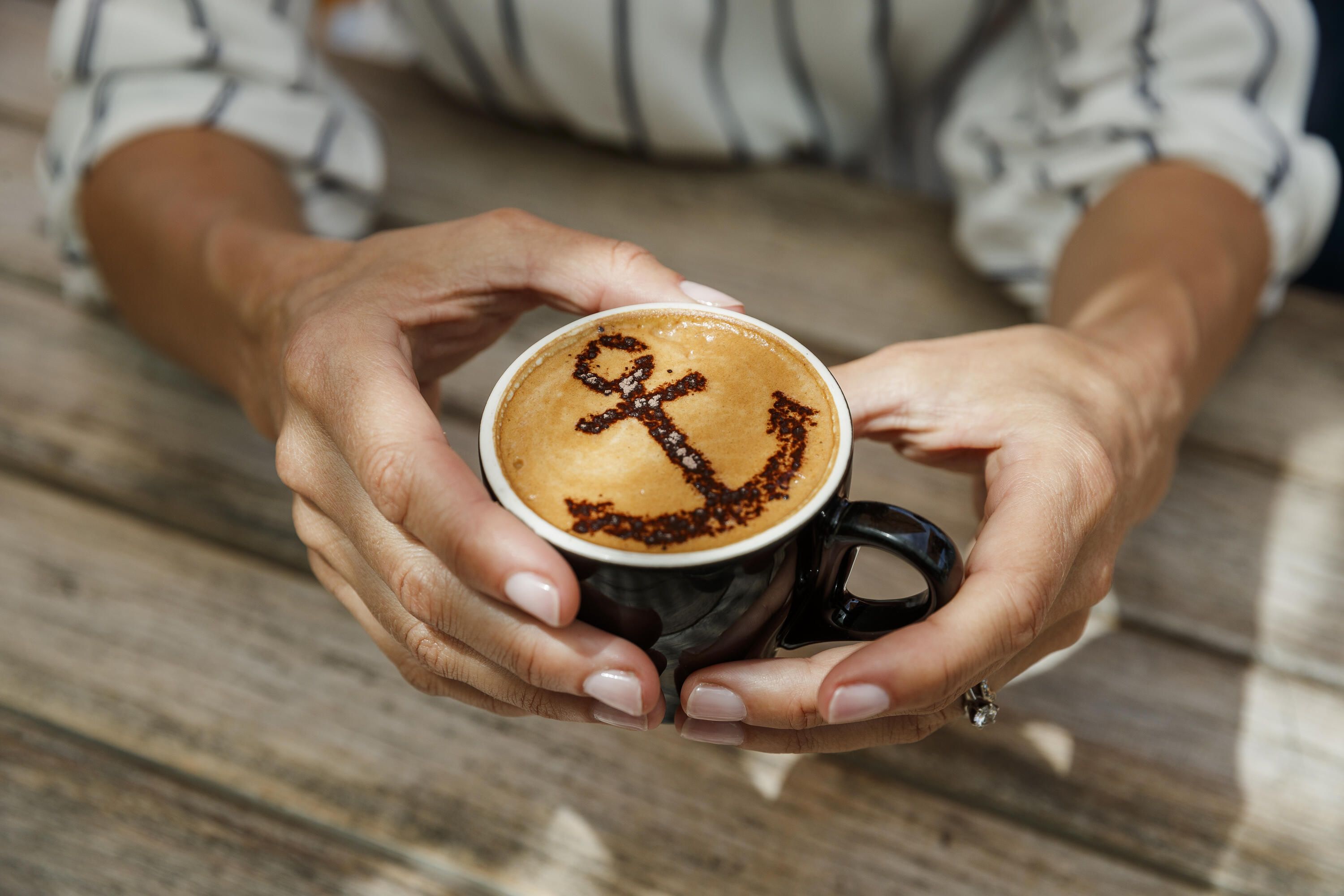 Close up photo showing hands holding a coffee with the shape of an Anchor on it.