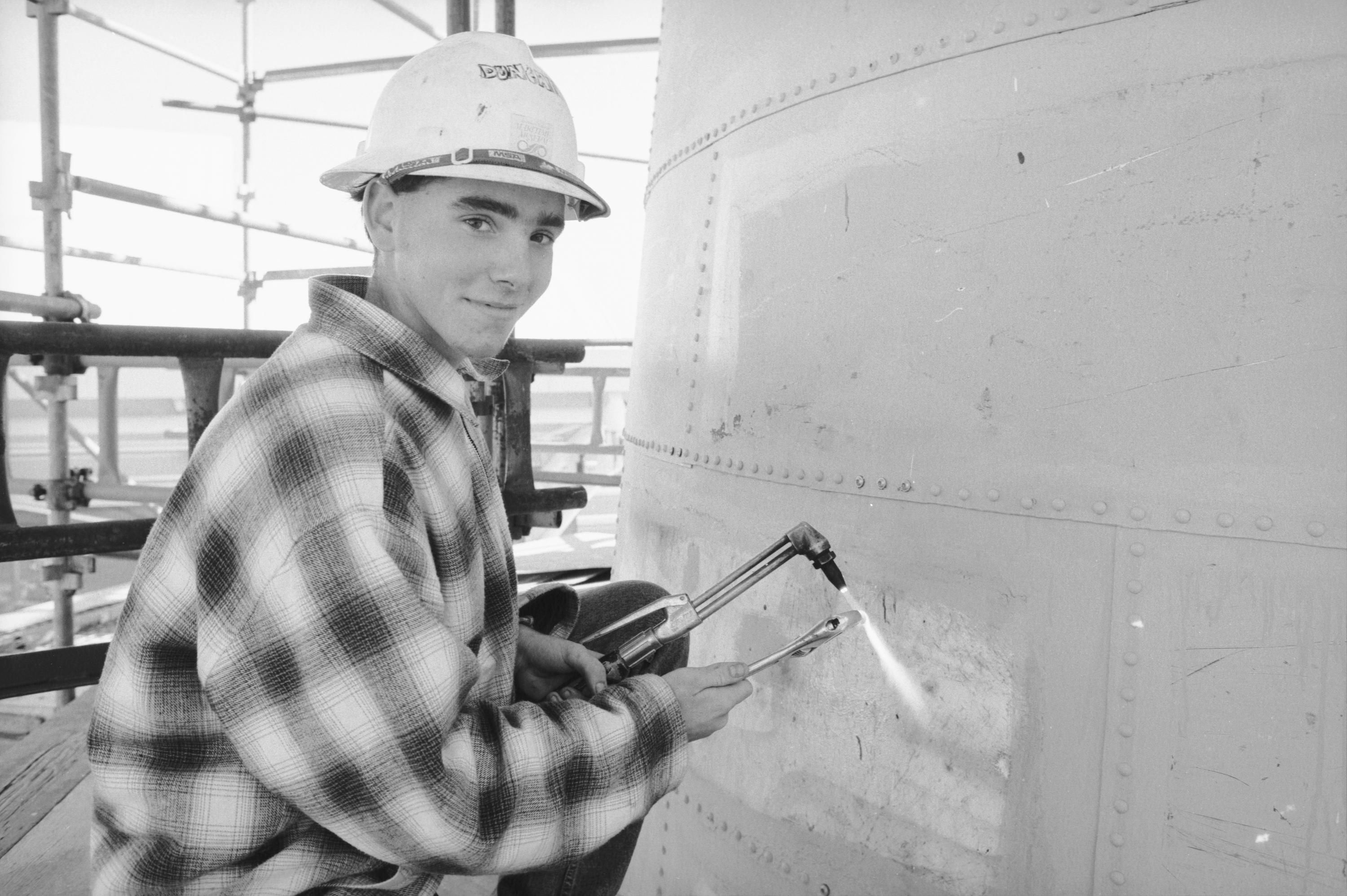 A portrait of a man working on a lighthouse, holding a blow torch and wrench. 