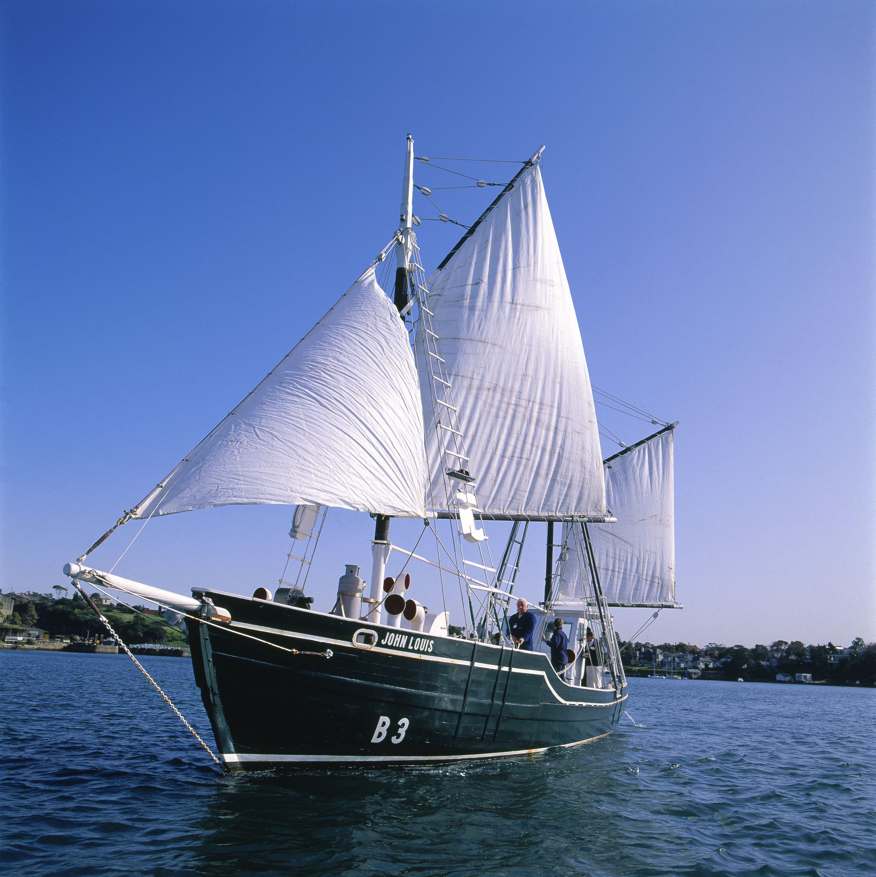 Photo of a boat with a dark green hull and white sails on blue water with a clear blue sky. 