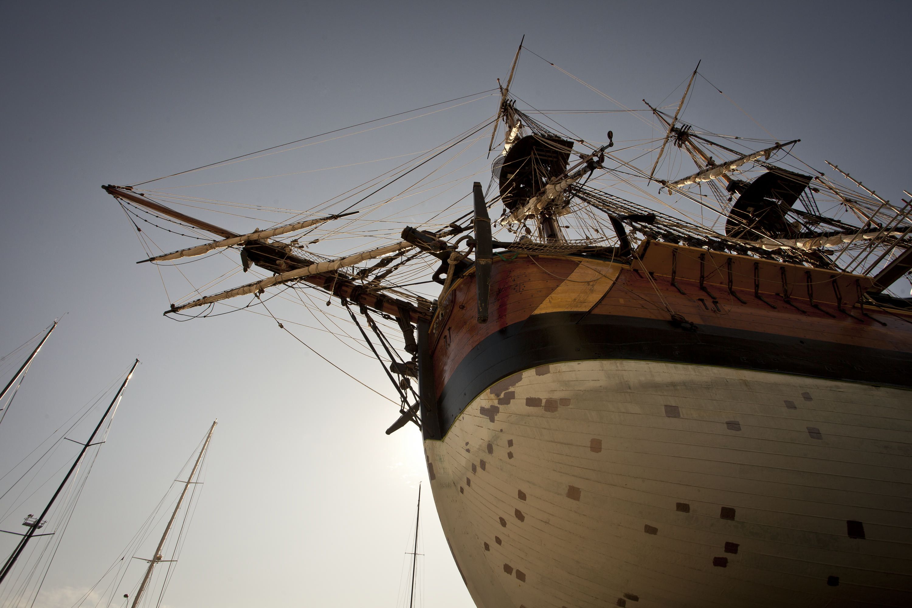 Photo taken upwards showing the hull of a tall ship and the masts and rigging (ropes) above. The sun is behind the ship giving the sky a soft glow. 