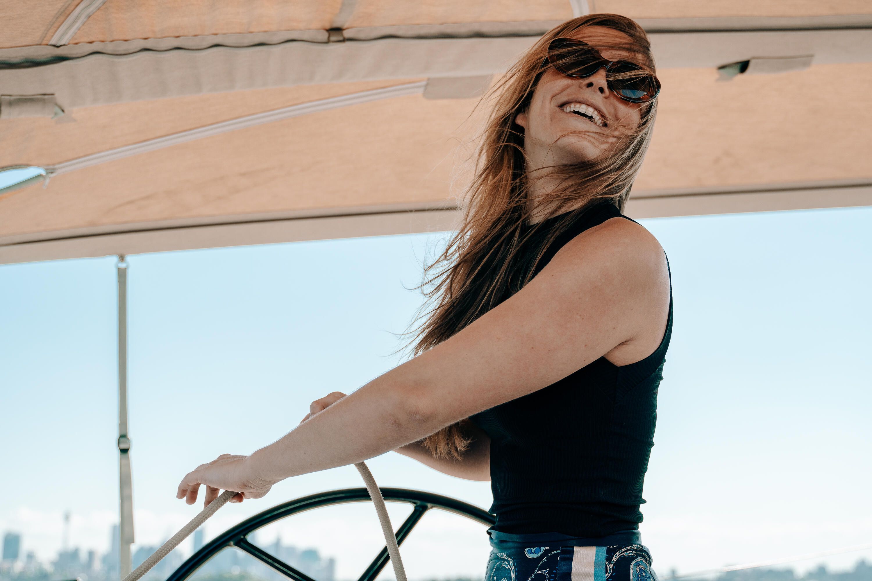 A woman wearing sunglasses and a black, sleevless top is smiling while holding a large steering wheel. The wind is blowing in her long hair. 