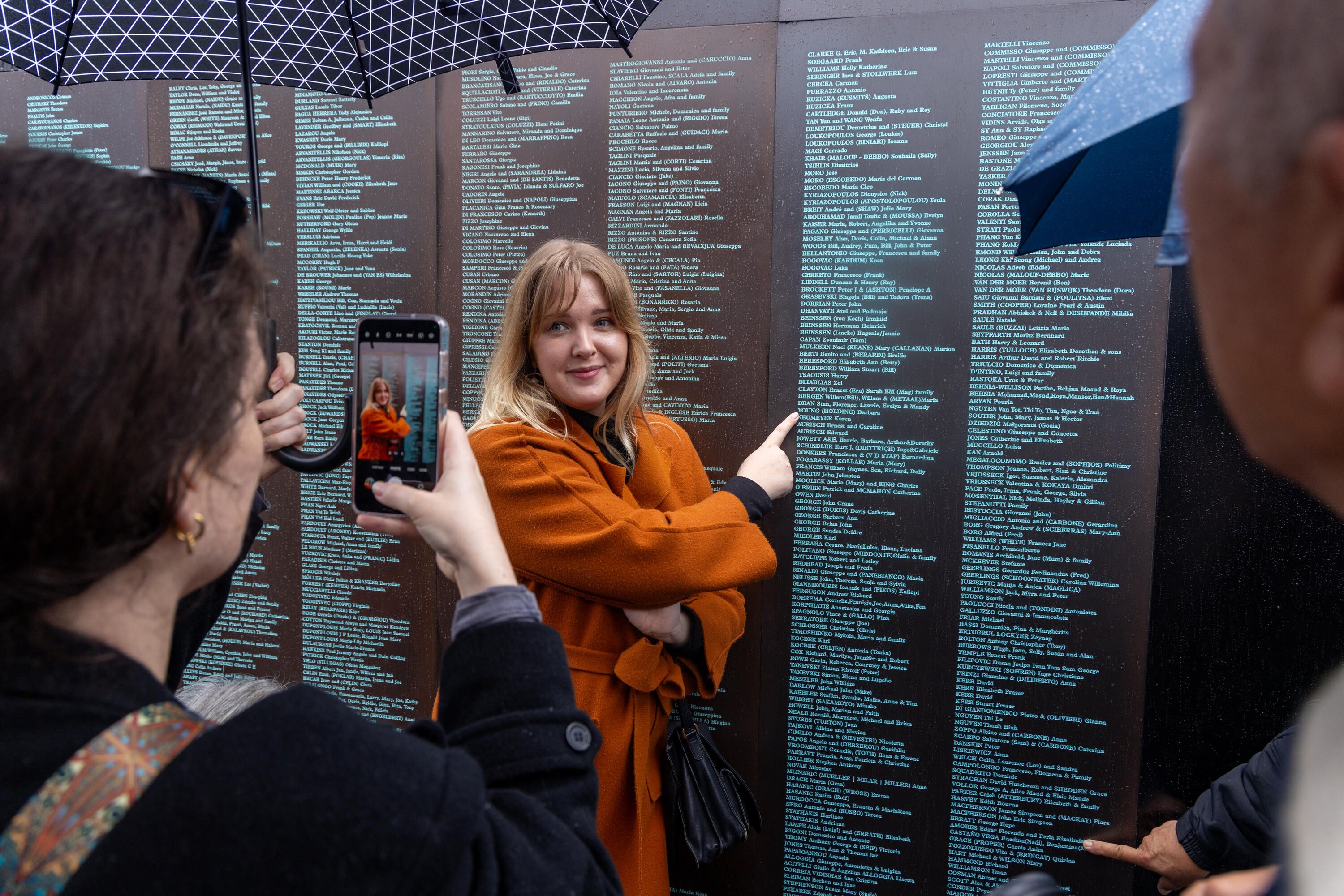 Photo of someone taking a photo of a lady in an orange coat pointing at a names on a dark, metal wall covered with small, green engravings.