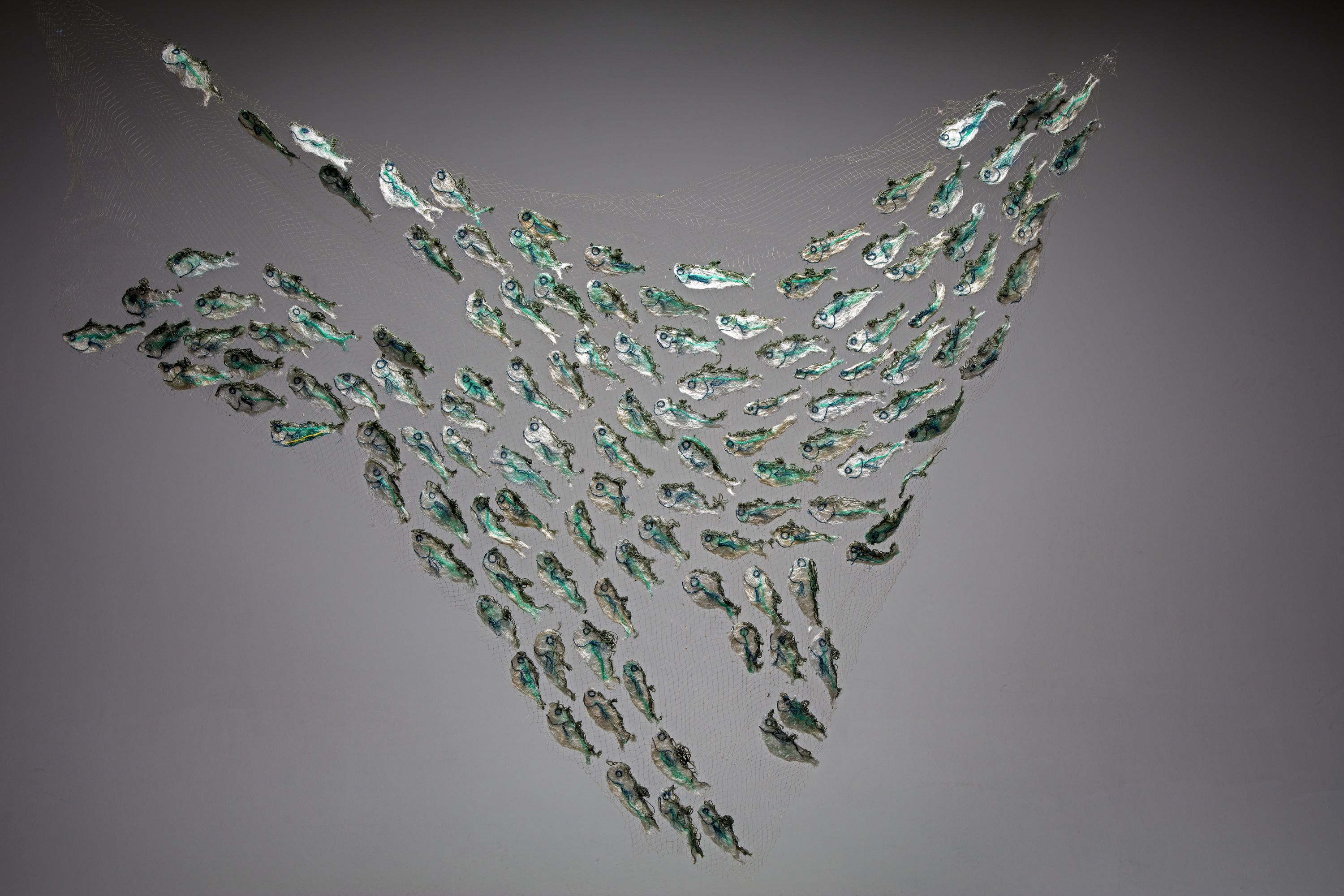 A sculpture of a school of sardines, all swimming in one direction but form various patterns. The backing is a clear net, with the sardines being white, teal green and black in colour.