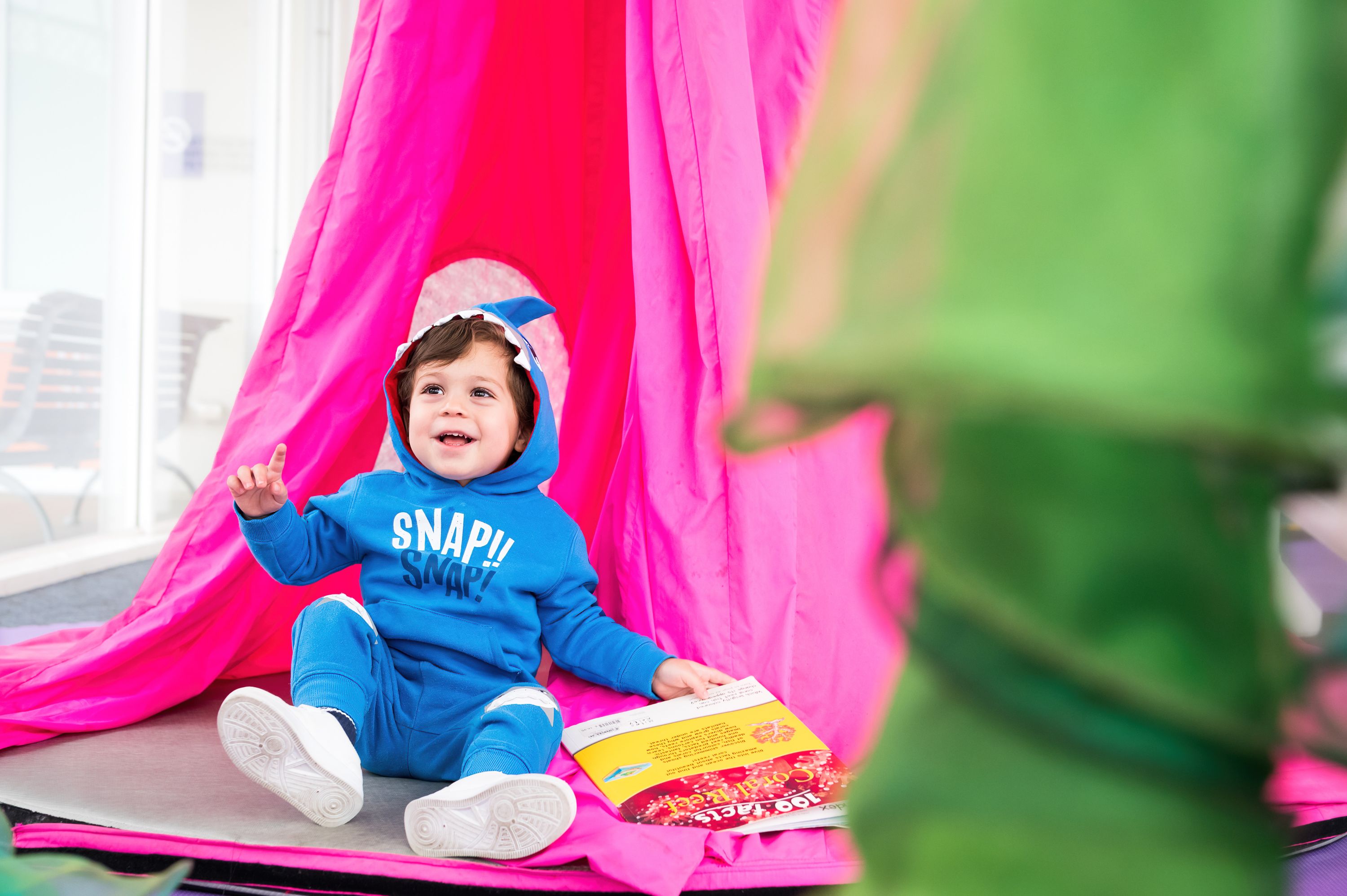 Photo of smiling boy wearing a blue hoodie with shark teeth, siting in front of a bright pink tent, holding a book.