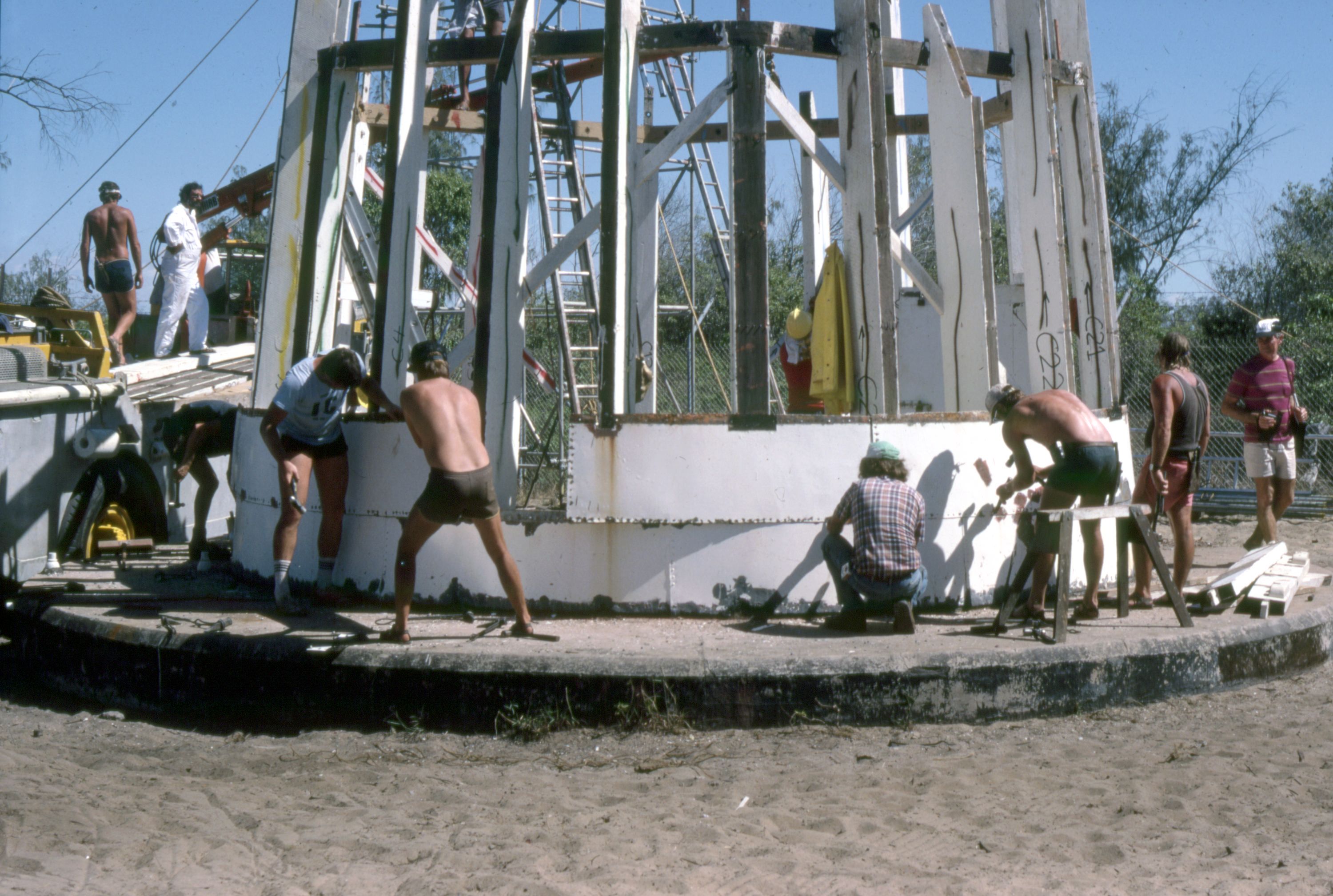 Photo showing a large group of men taking off the curved metal sheeting from the outside of a lighthouse. 