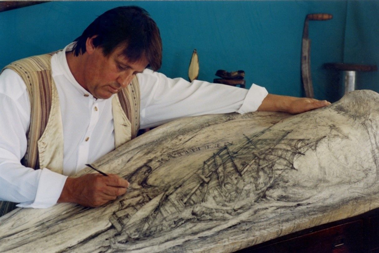 Photo of a male artist wearing white and cream, working on an extremely large piece of bone.