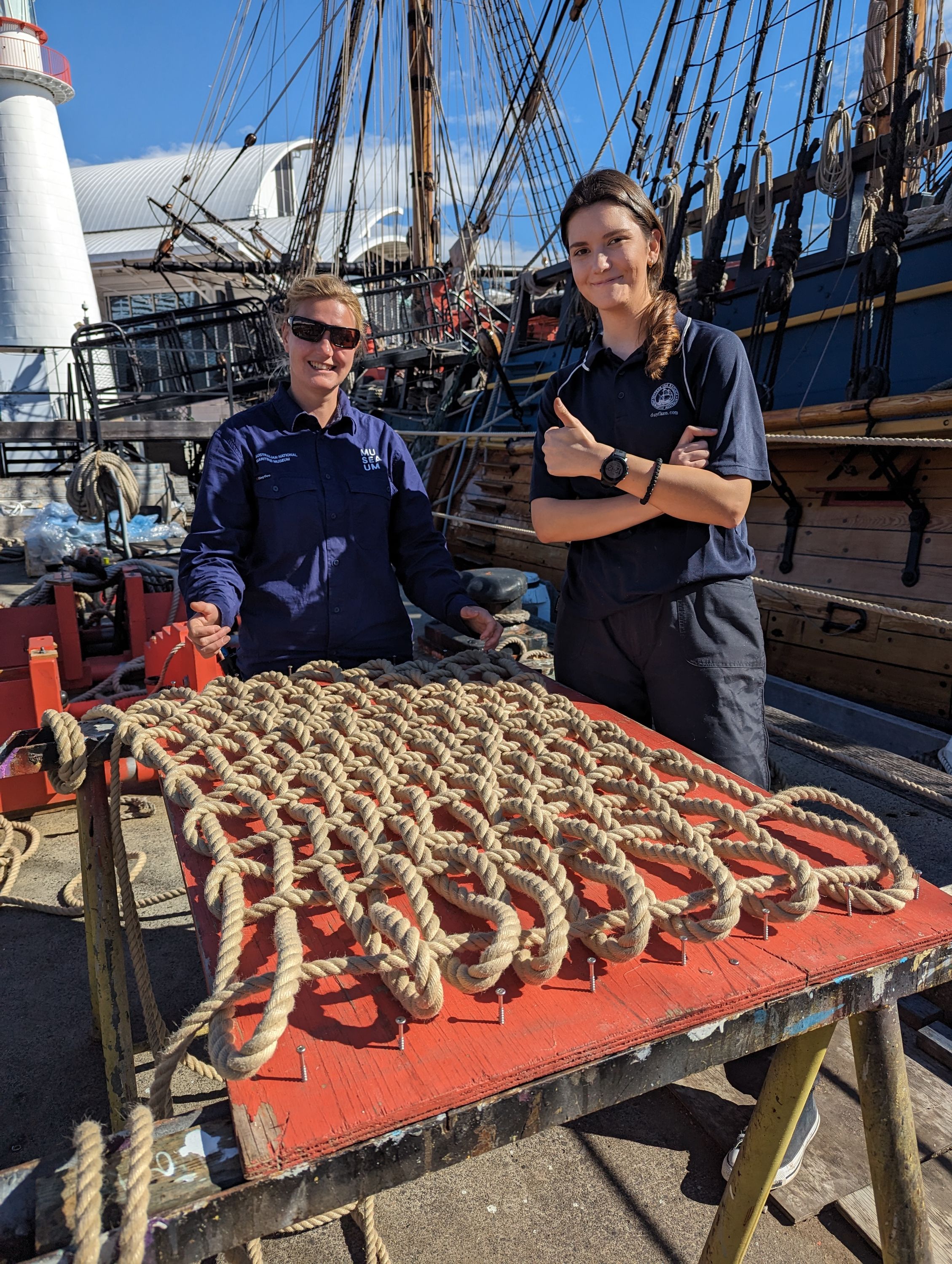 Photo of two women in navy museum uniform, standing behind woven ropework.