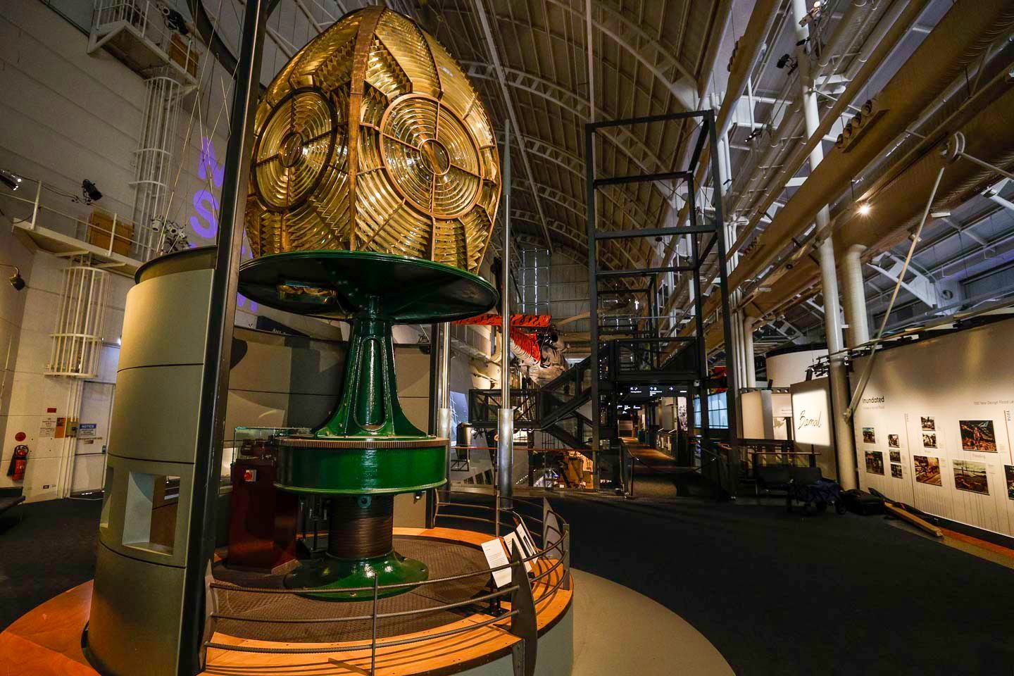Photo of inside a museum gallery. The gallery features a large lighthouse lantern in the middle of the room and a display of images on the wall behind it.