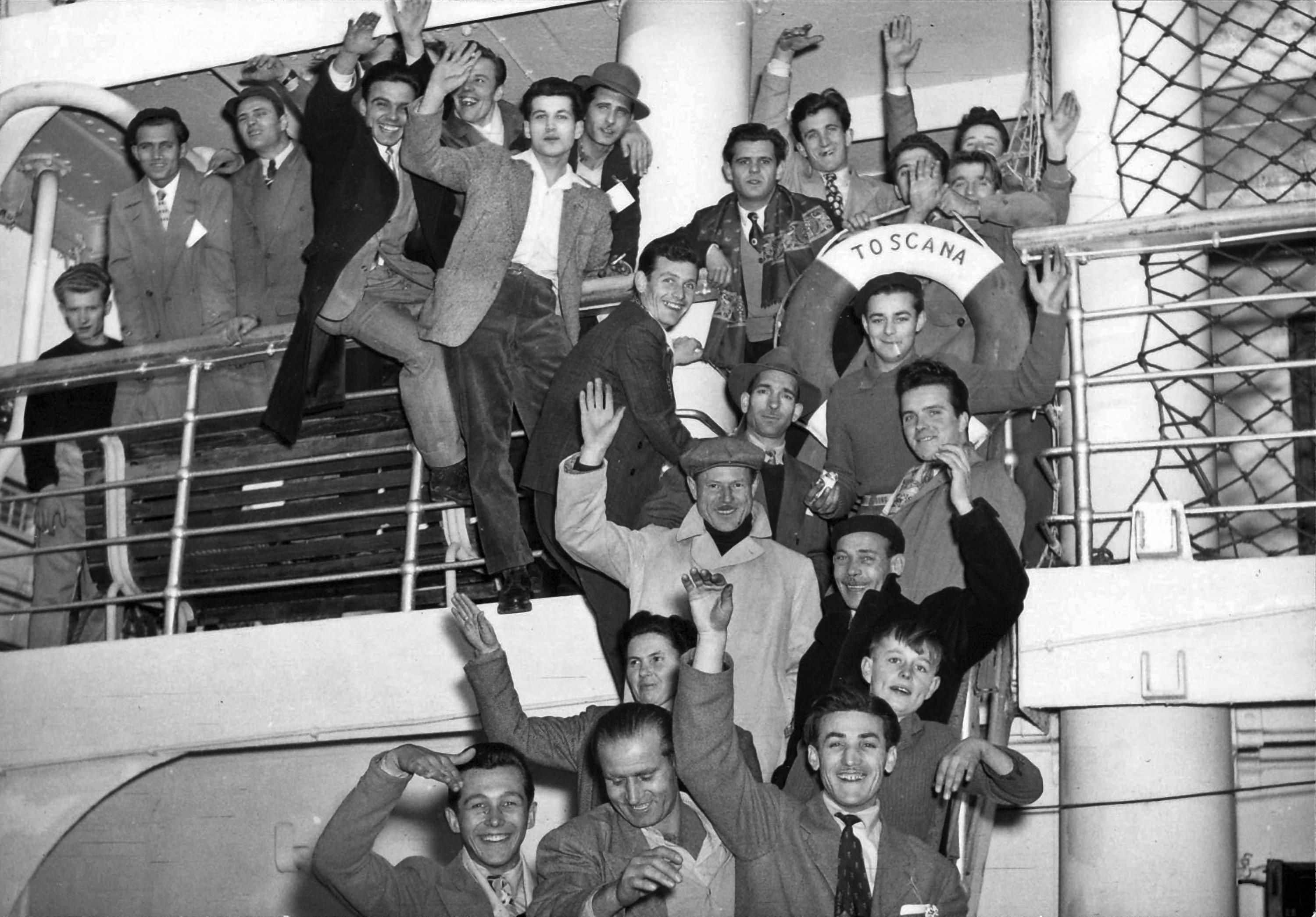 Black and white photo of about two dozen male adults in heavy clothes on deck and companionway of a ship looking and waving at camera