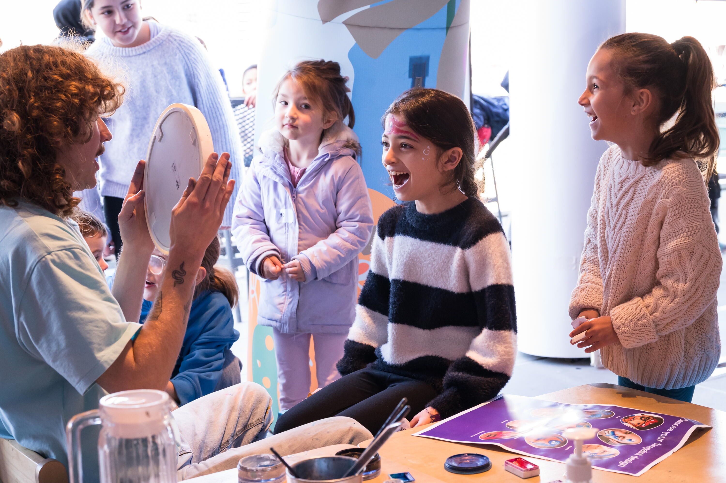 girl looking at herself in a mirror which is being held up by a museum staff member who has painted an octopus on her face. There are 2 other girls looking on.