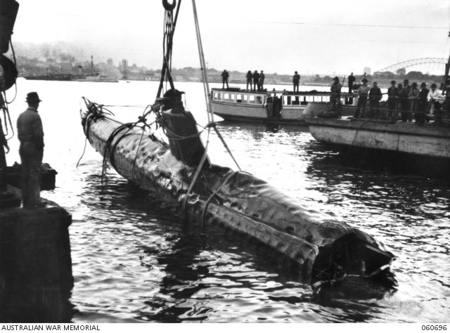Black and white photo showing a small, damaged submarine being lifted out of the water, presumably by a crane.