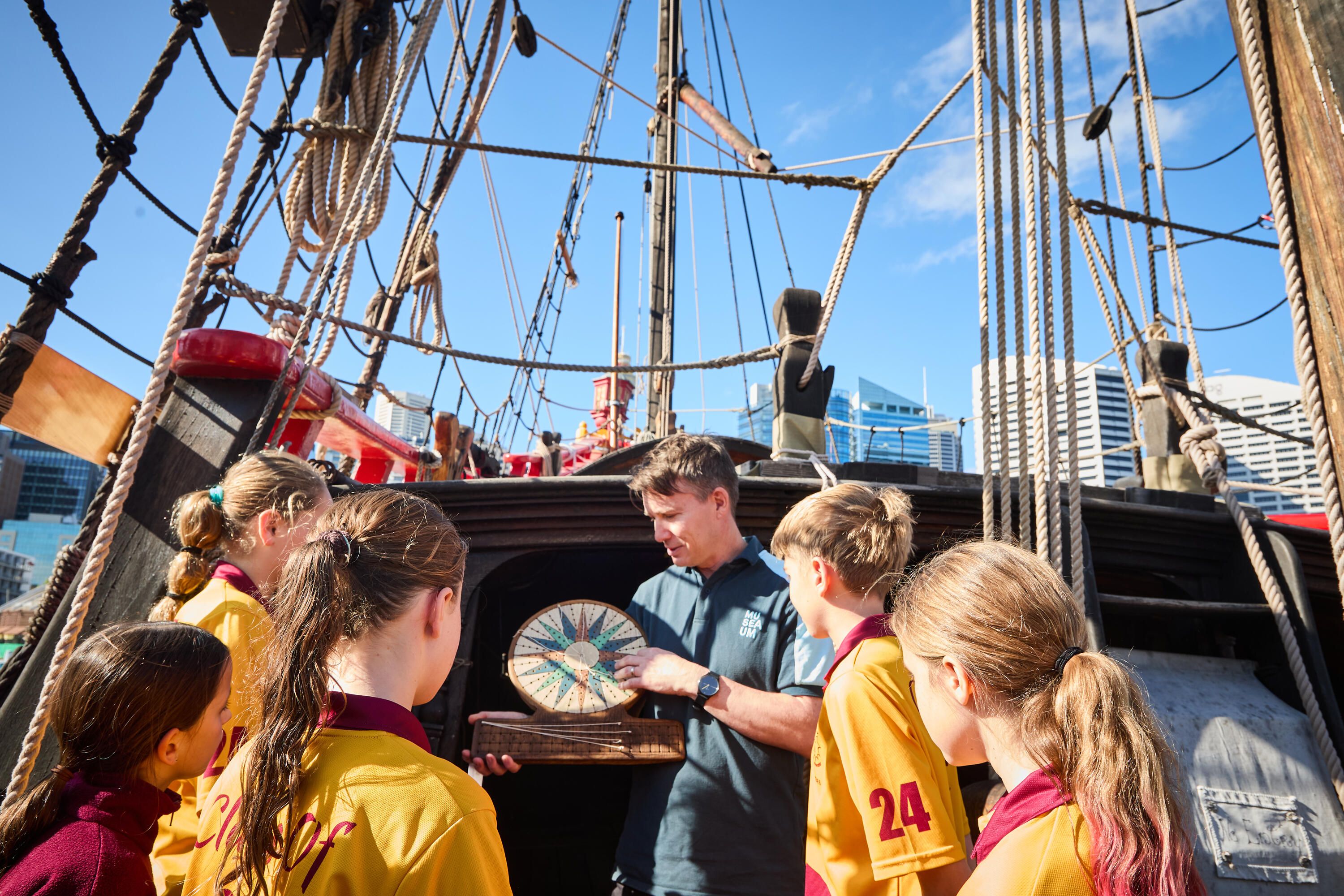 Group of school students and a museum educator standing outside in the sun, onboard a wooden tallship.