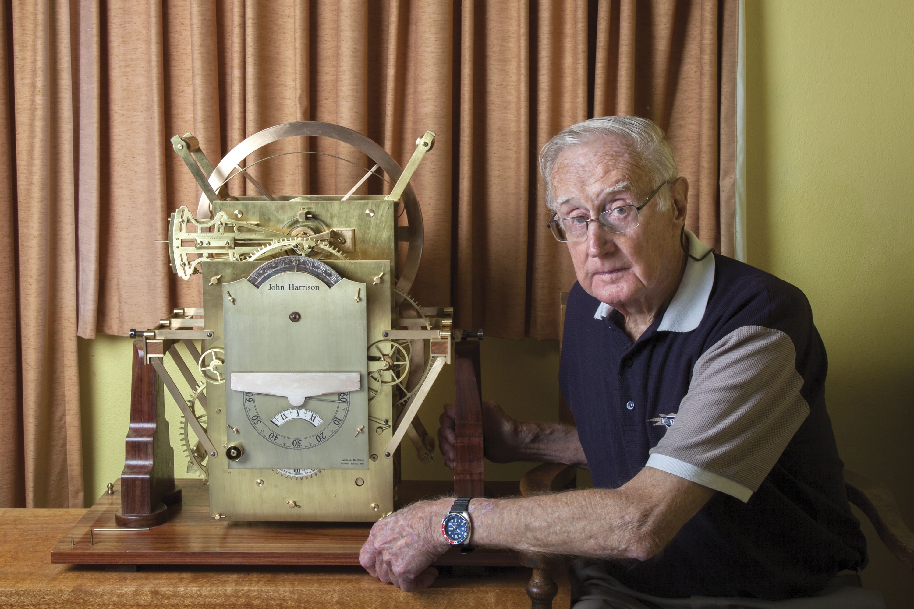 Photograph showing an older man sitting next to a large, elaborate clock. 