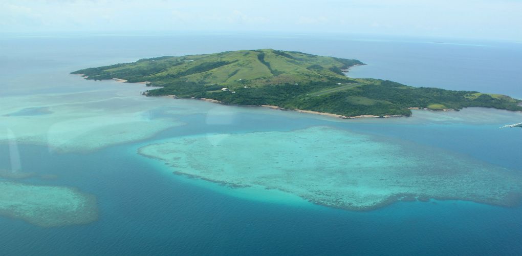Photo taken from the air of a green island covered in vegetation, surrounded by aqua blue water with submerged reefs.