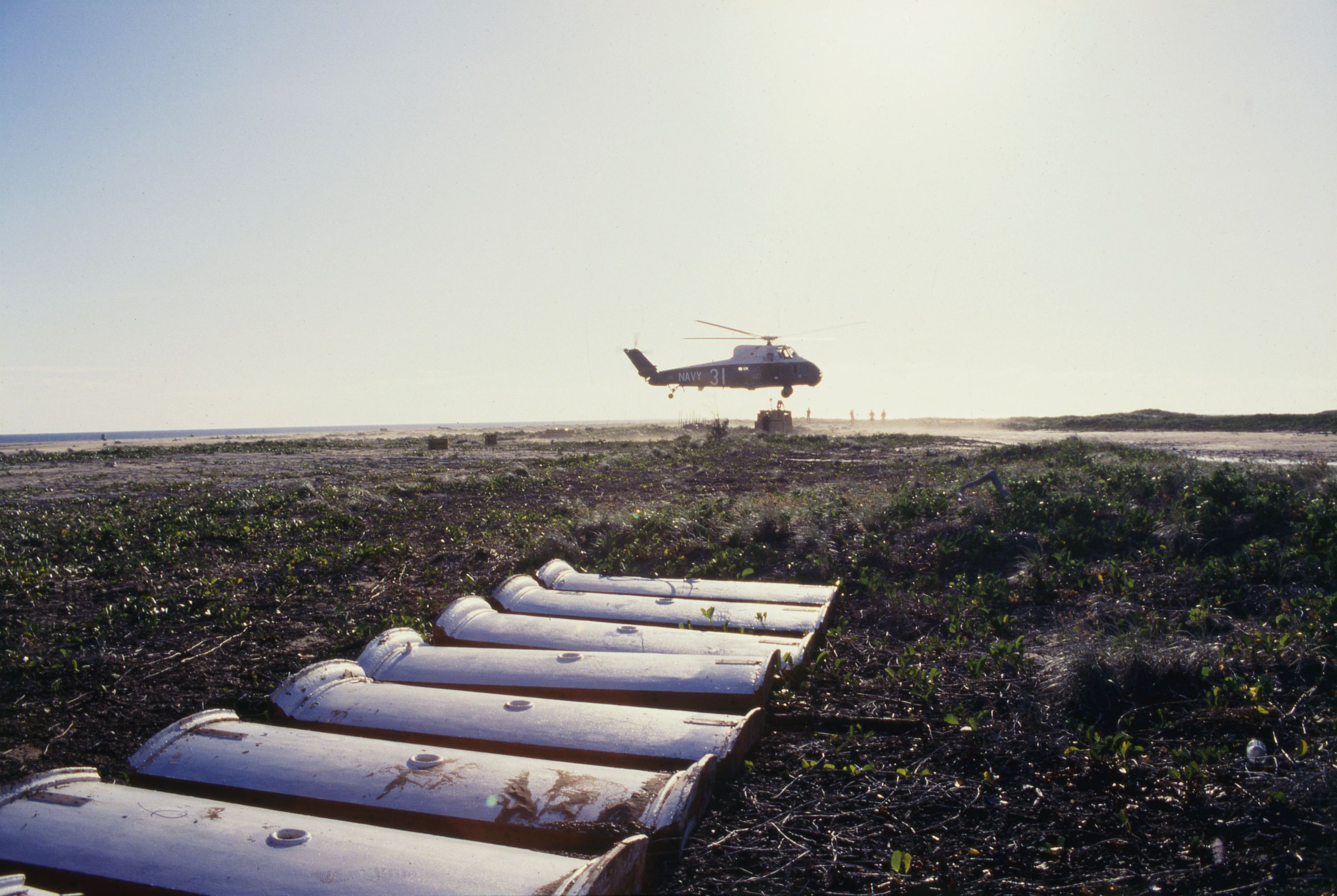 Photo showing a helicopter lifting cargo in a net. Pieces of curved metal are lined up in the foreground. 