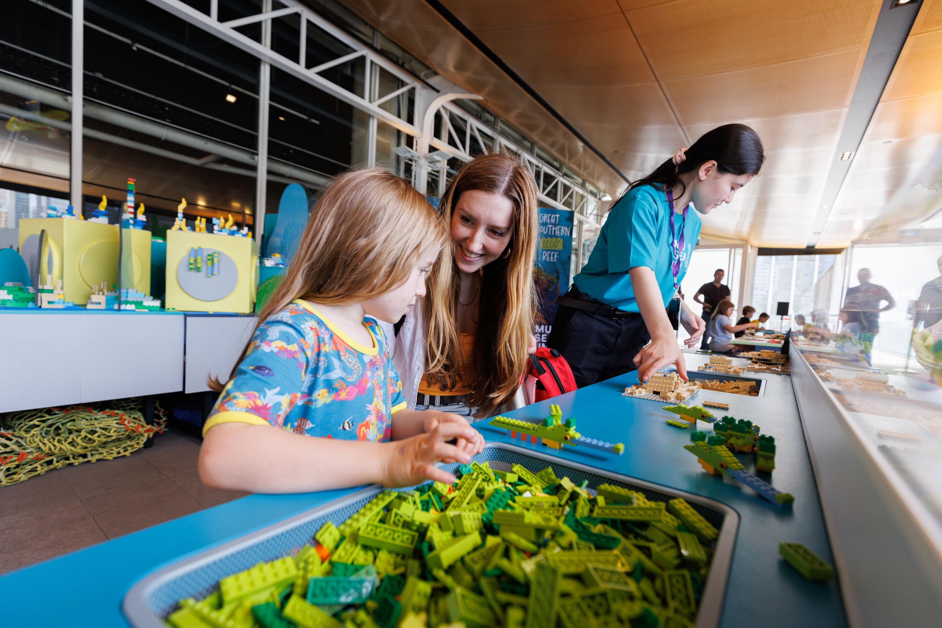 Photograph of a young girl and an adult woman seated at a table making lego together. A museum staff member is in the background.
