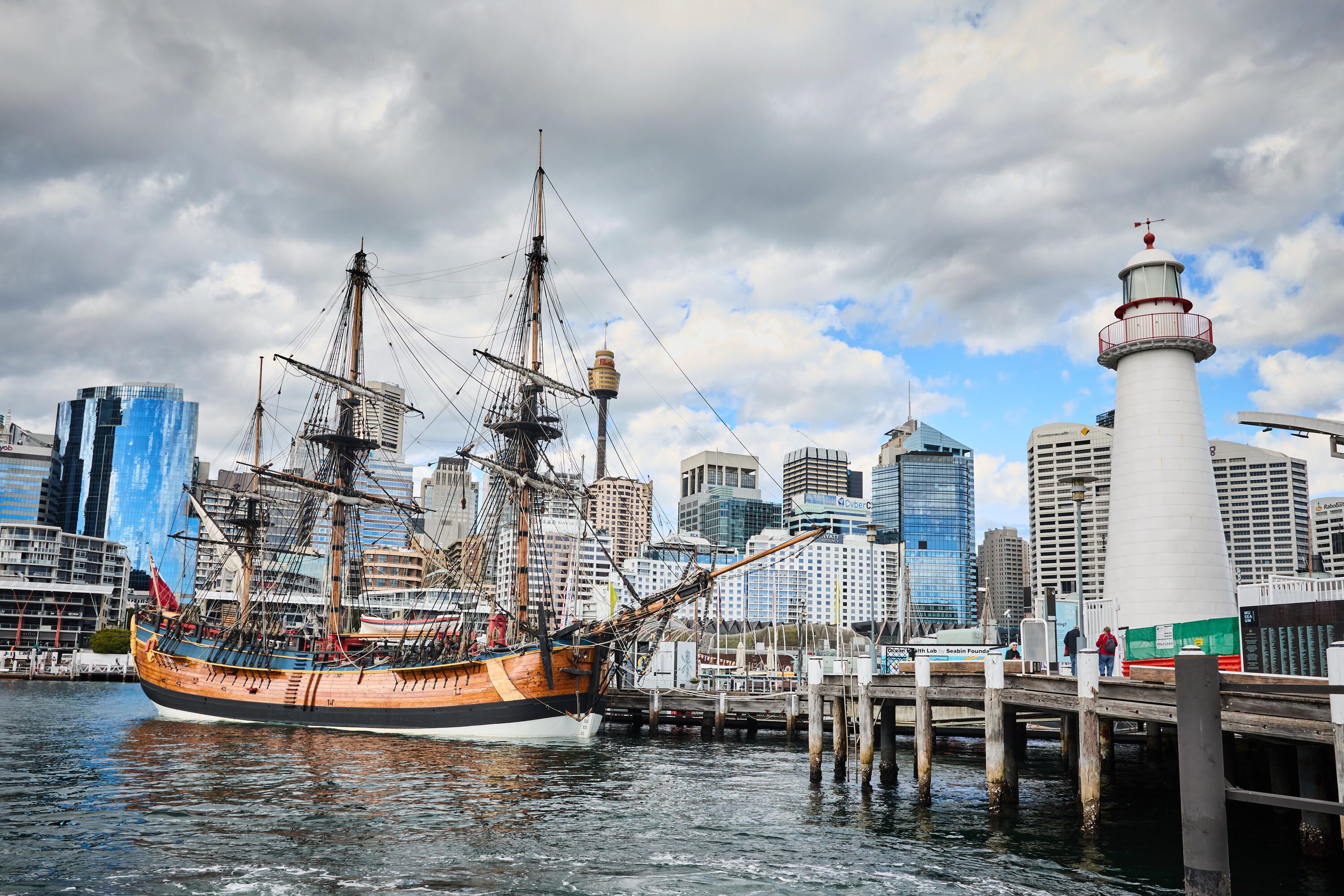 Photo showing a wooden tall ship next to a wharf and white lighthouse. 