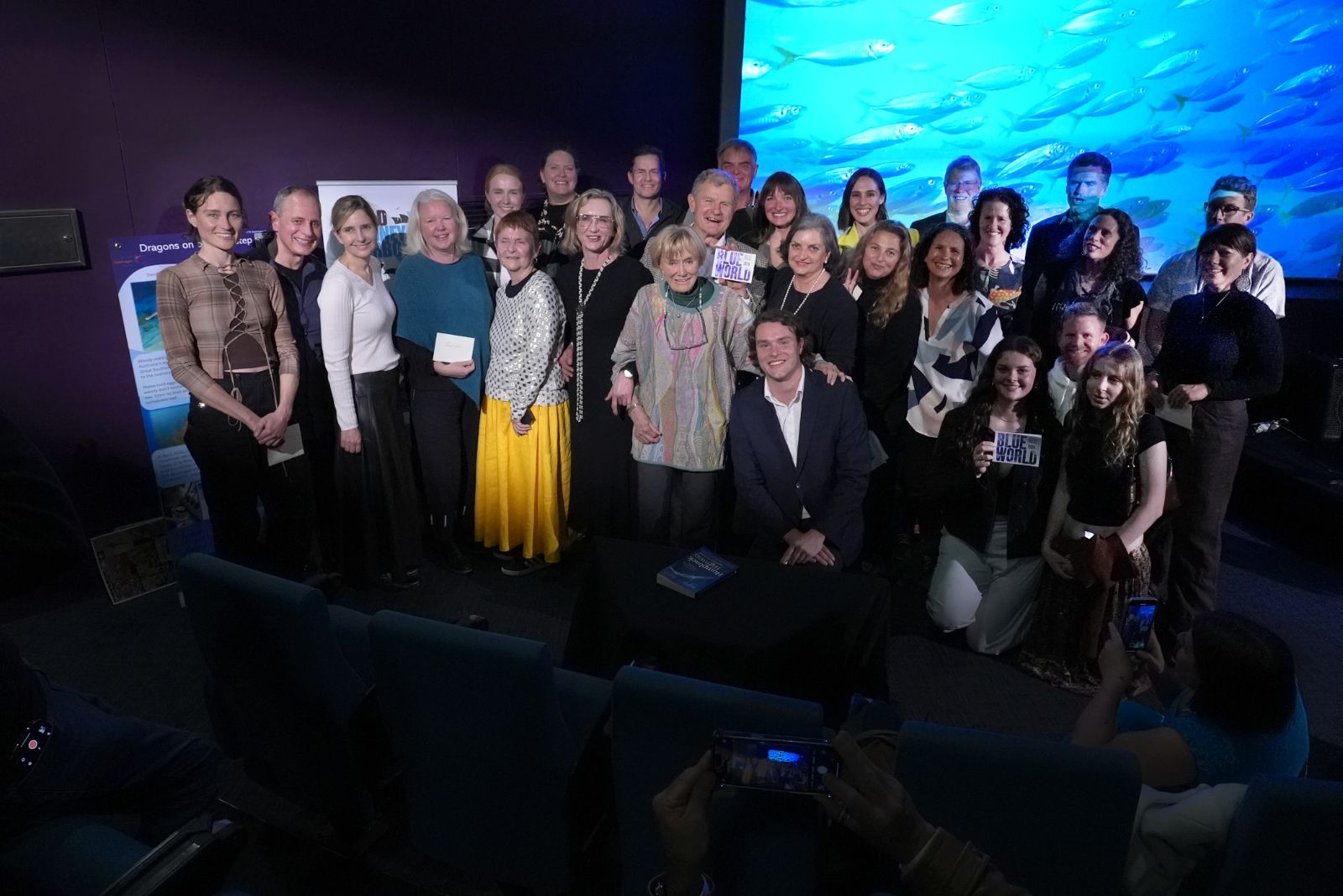 Photo in a dark theatre with a large group of people posing for the photo in front of the screen.
