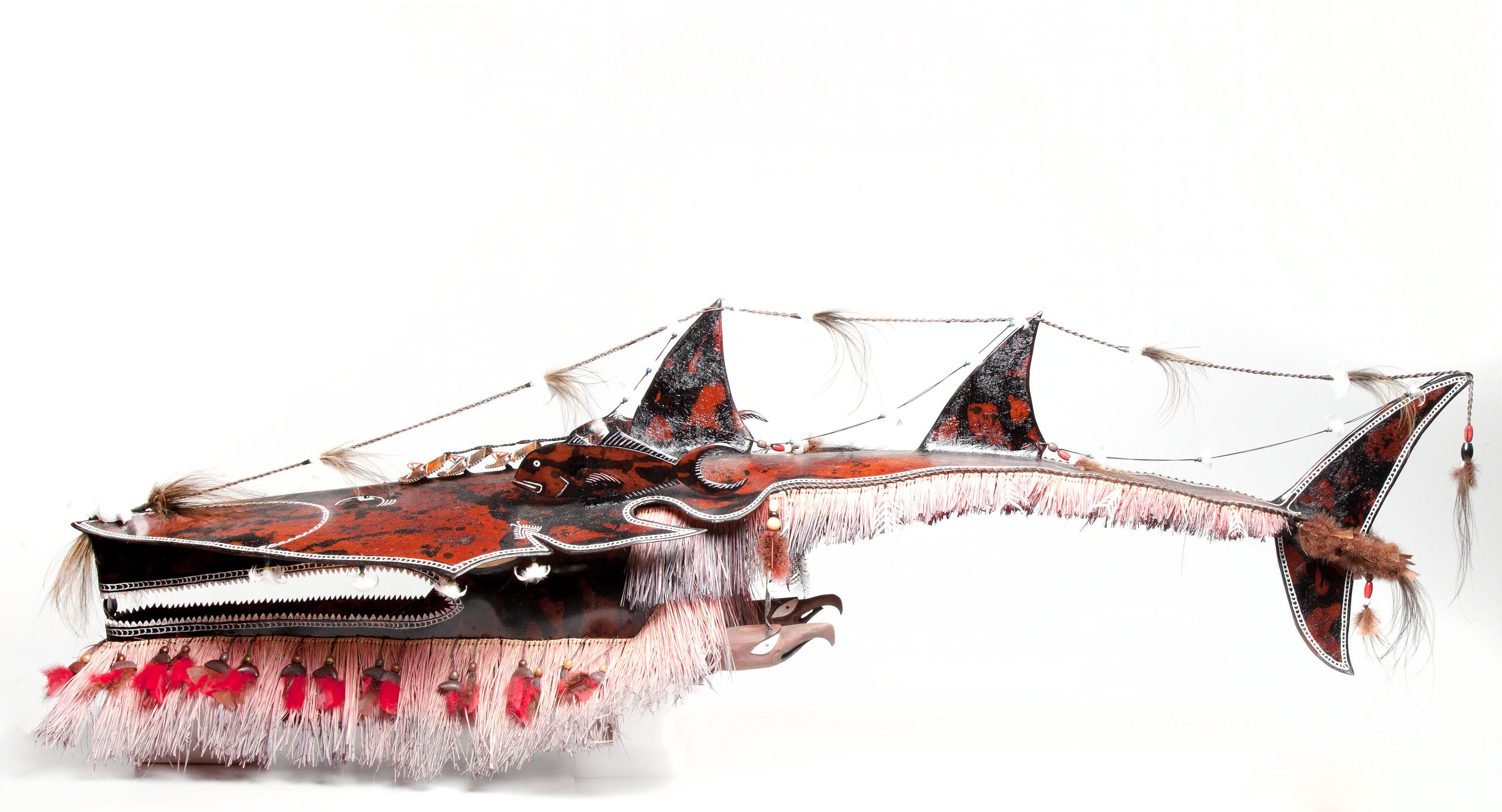 Sculpture of a shark with a semi open mouth and red fibre hanging from under the head and tail. Inside the mouth, a human like mask. There are also four smaller masks on a line on top of the shark’s head. In between the head and the tail, a bird's head facing the tail on each side of the shark. There is a string with feathers going from the nose to the tip of the tail. The eyes are made of shell and inlaid onto the surface. The shark is heavily decorated with rope, feathers and shells.
