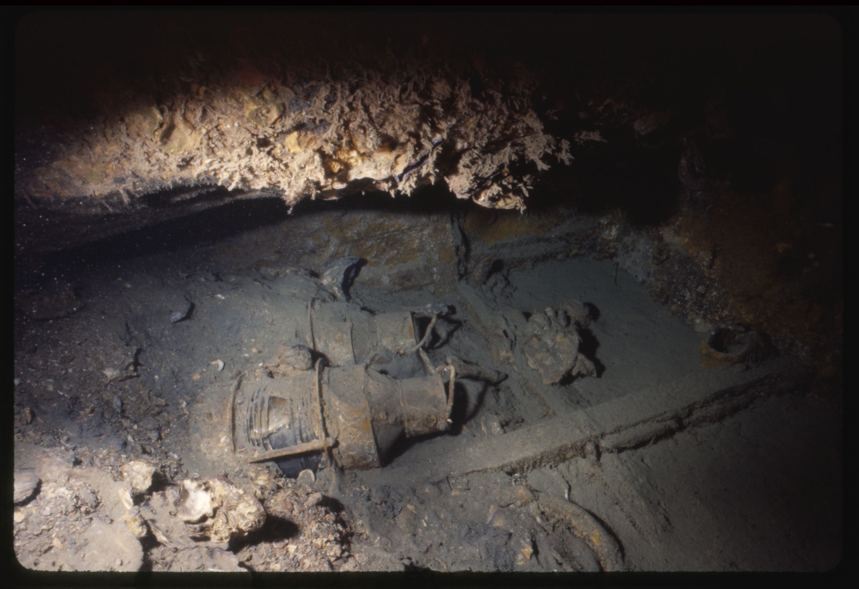 Photo taken underwater showing a shipwreck. Lanterns on their side are just visible and covered with sediment.  