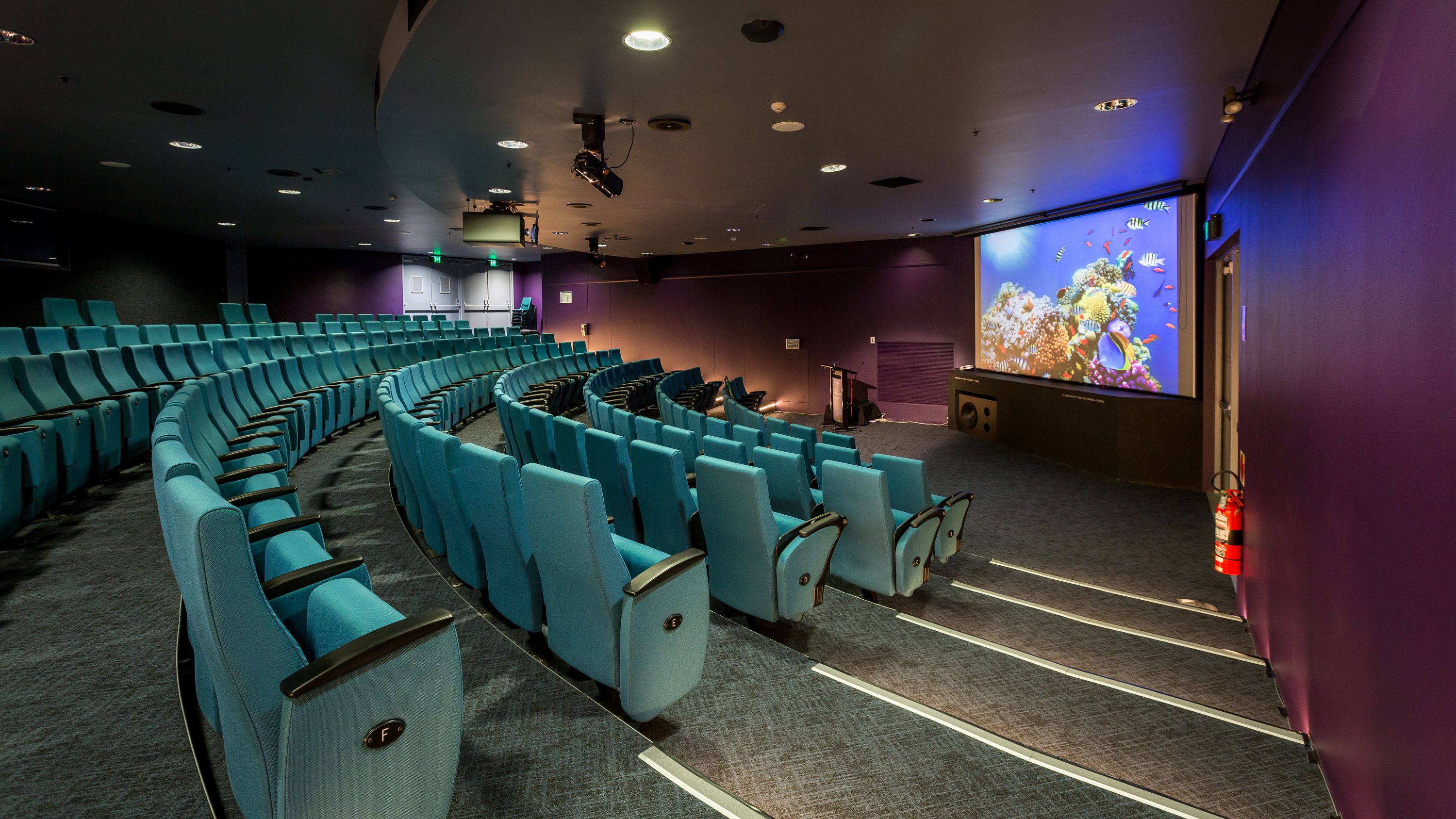 photo of an empty theatre with blue seats and a large screen at the front.