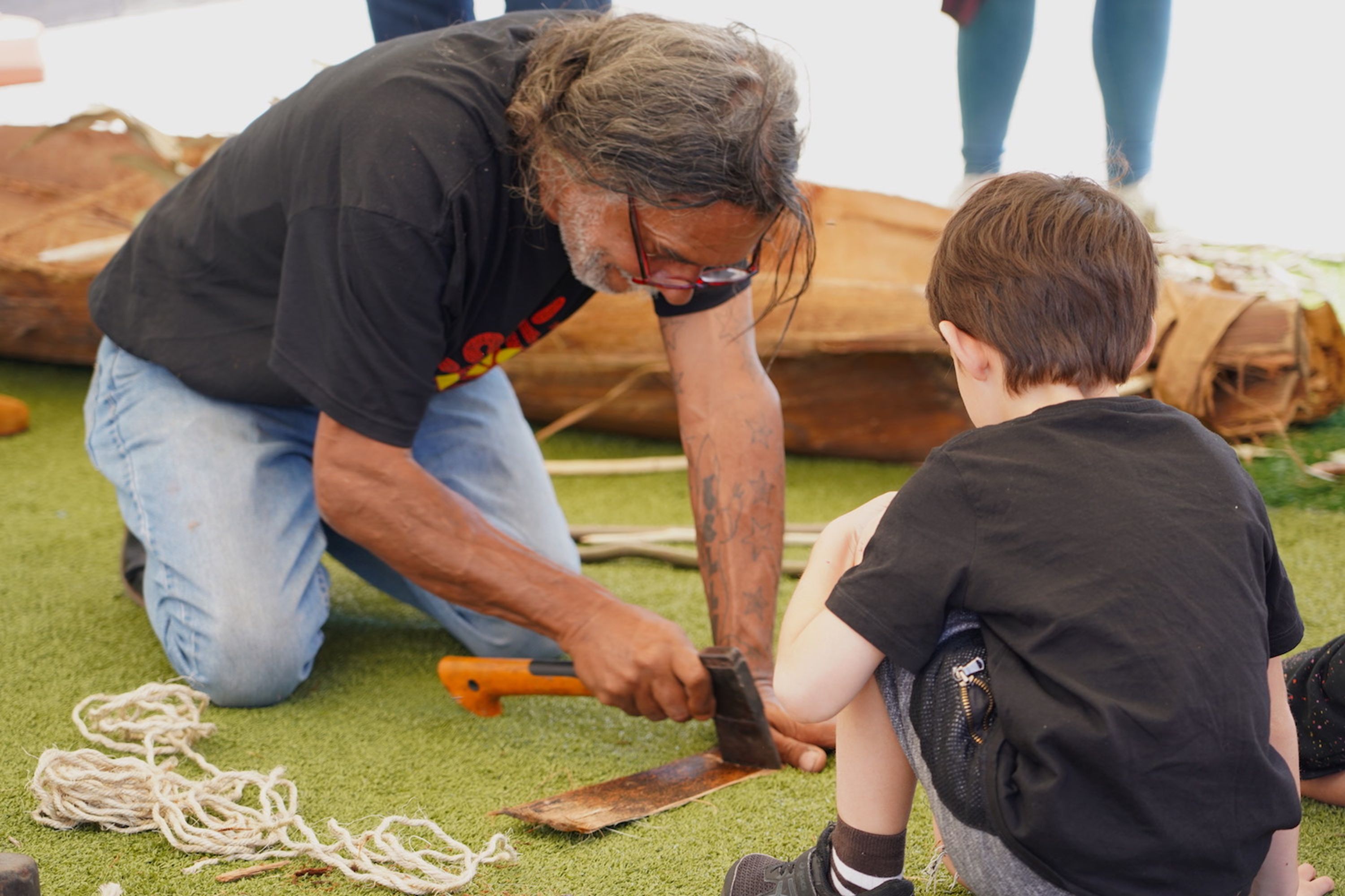 Man and a boy kneeling on the ground. The man us using a small axe to work on a piece of bark.
