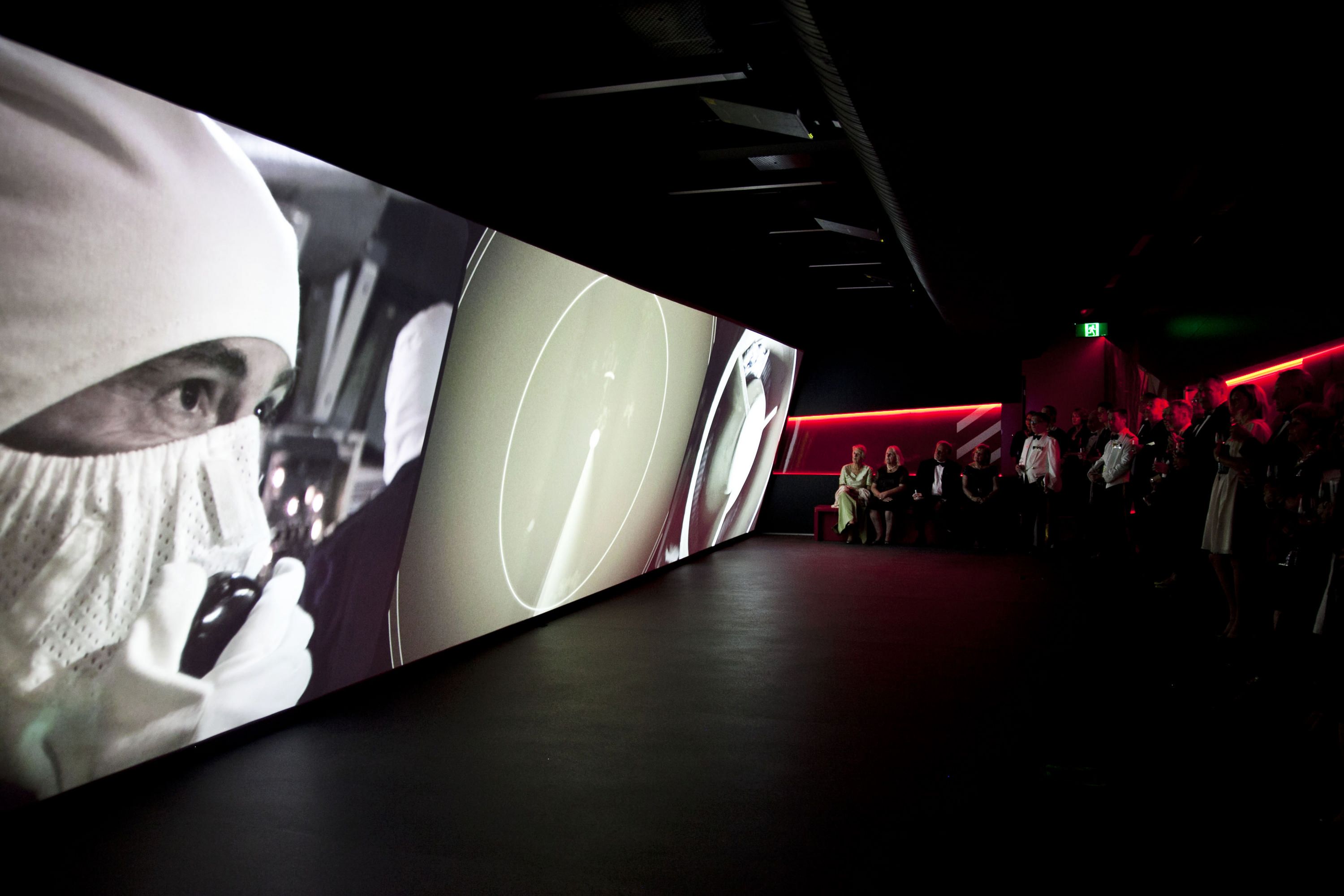 Photo showing a long, sloped screen showing a film depicting operations on the naval vessels, with a small crowd of people watching. 