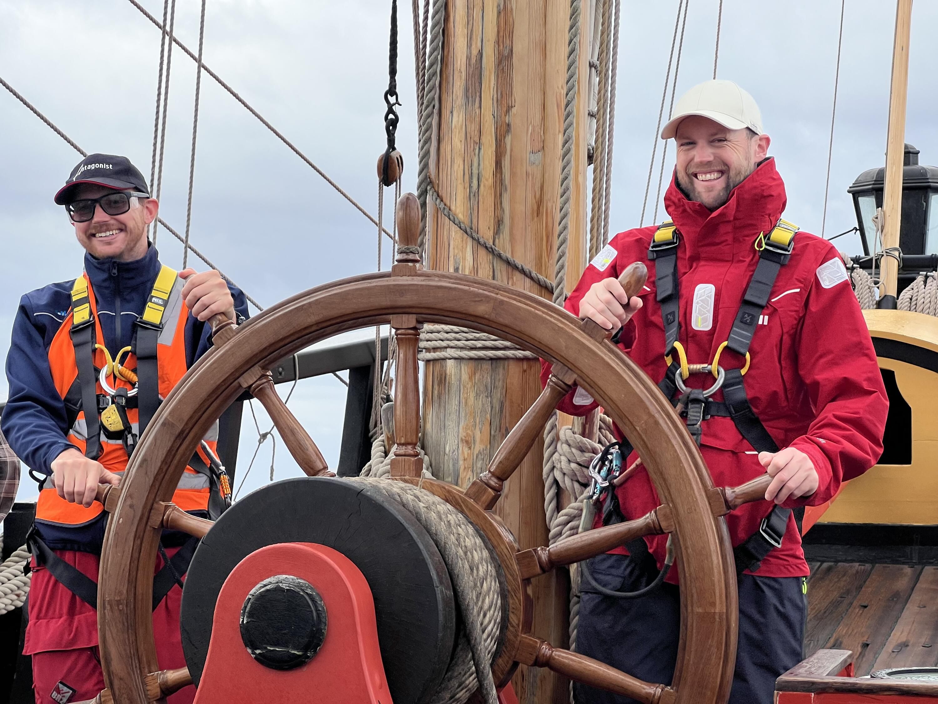 Photo showing 2 men wearing warm jackets, climbing harnesses and caps standing at a tall ship's wheel.