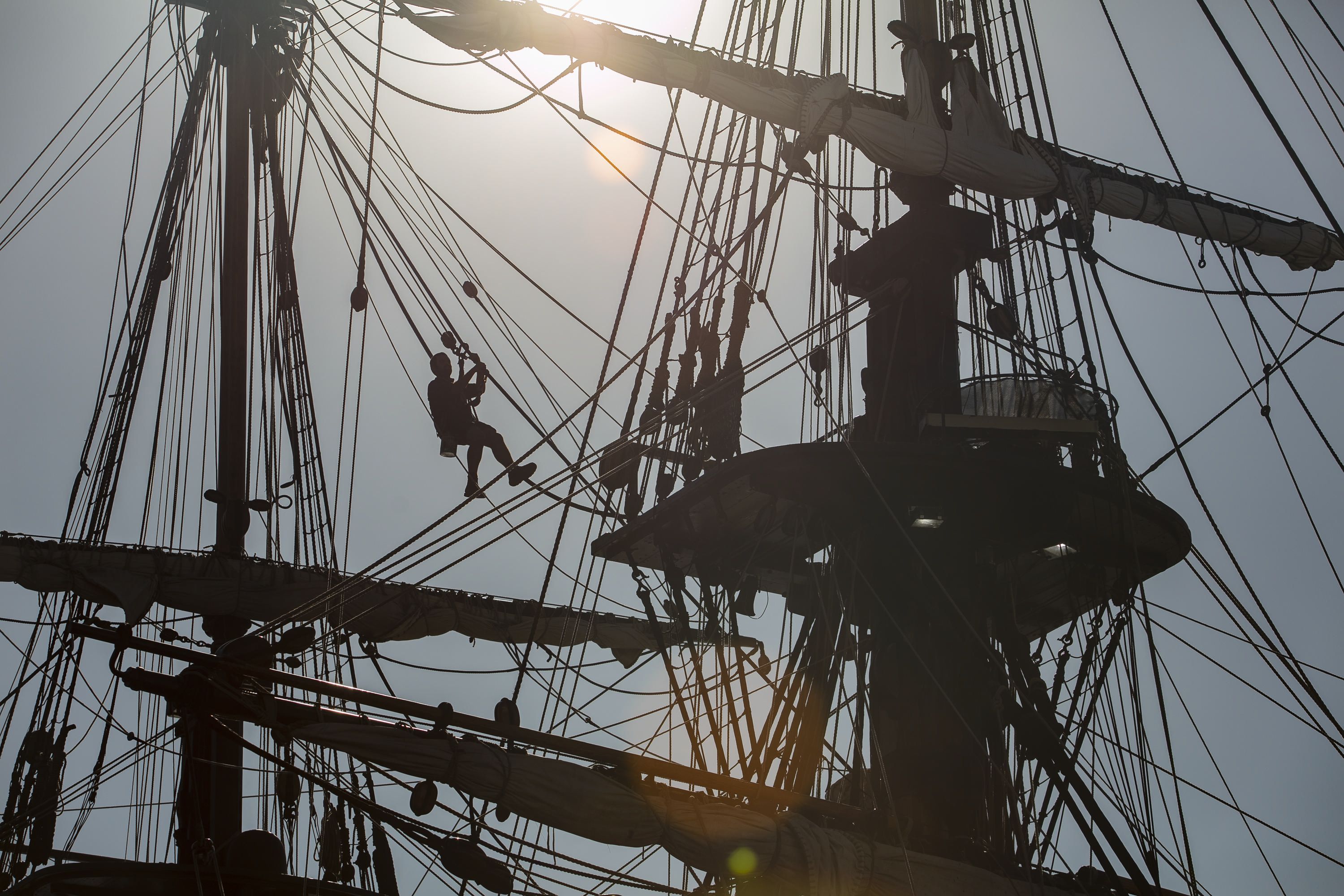 Photo of a silhouetted figure in a harness moving between the ropes between two masts of a tall ship. 