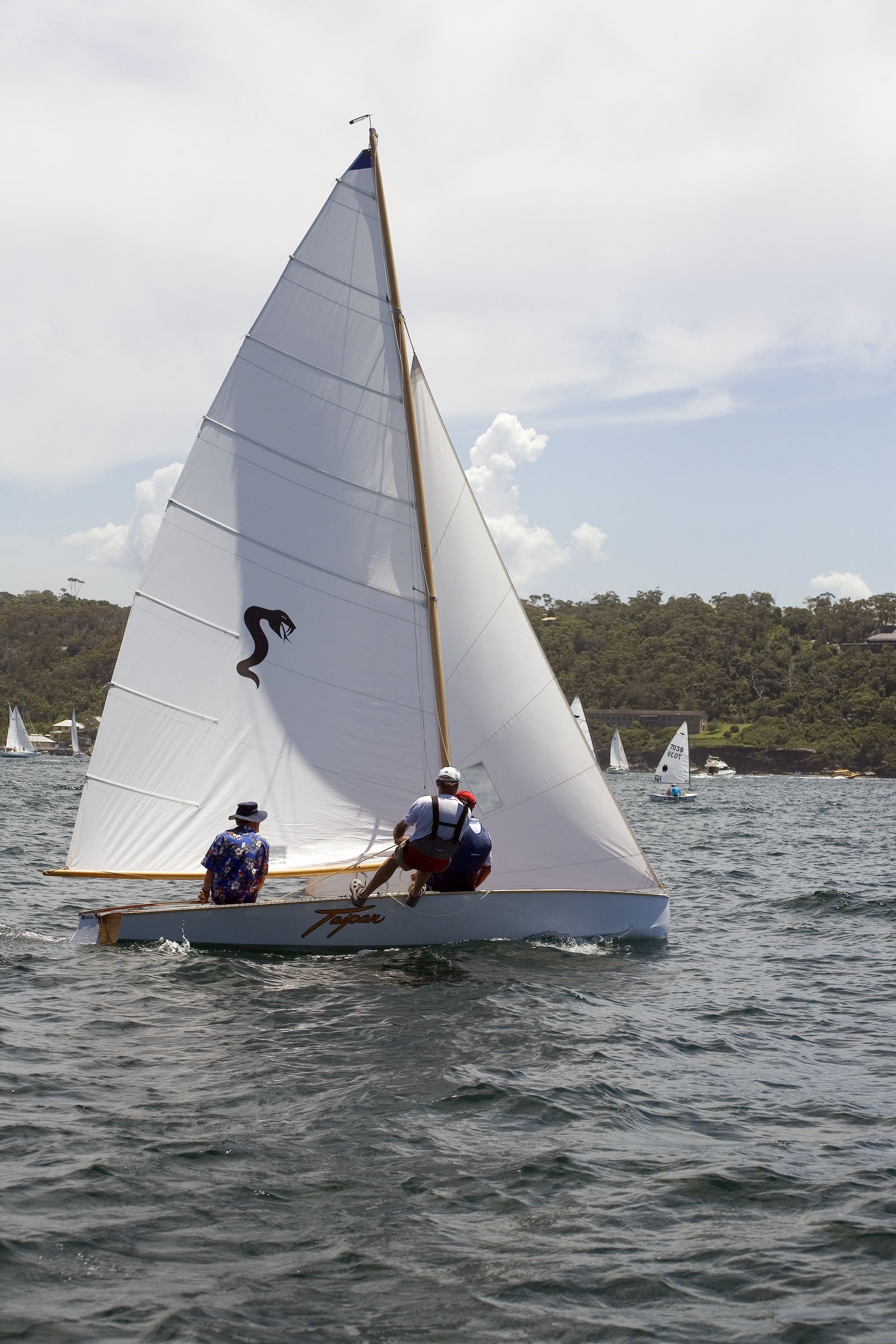 Photograph of a small sailing boat with 2 people sailing. The triangular sail is up and there are other boats in the background. 