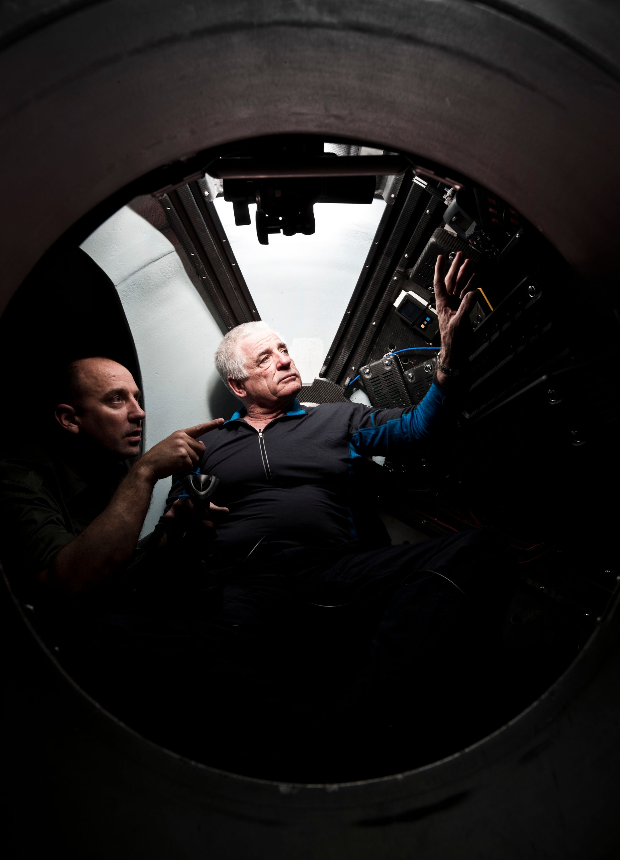 A man with white hair is seated in a sphere looking up and to the right of the image. A second man is just visable at the left of the image pointing to something on the right.