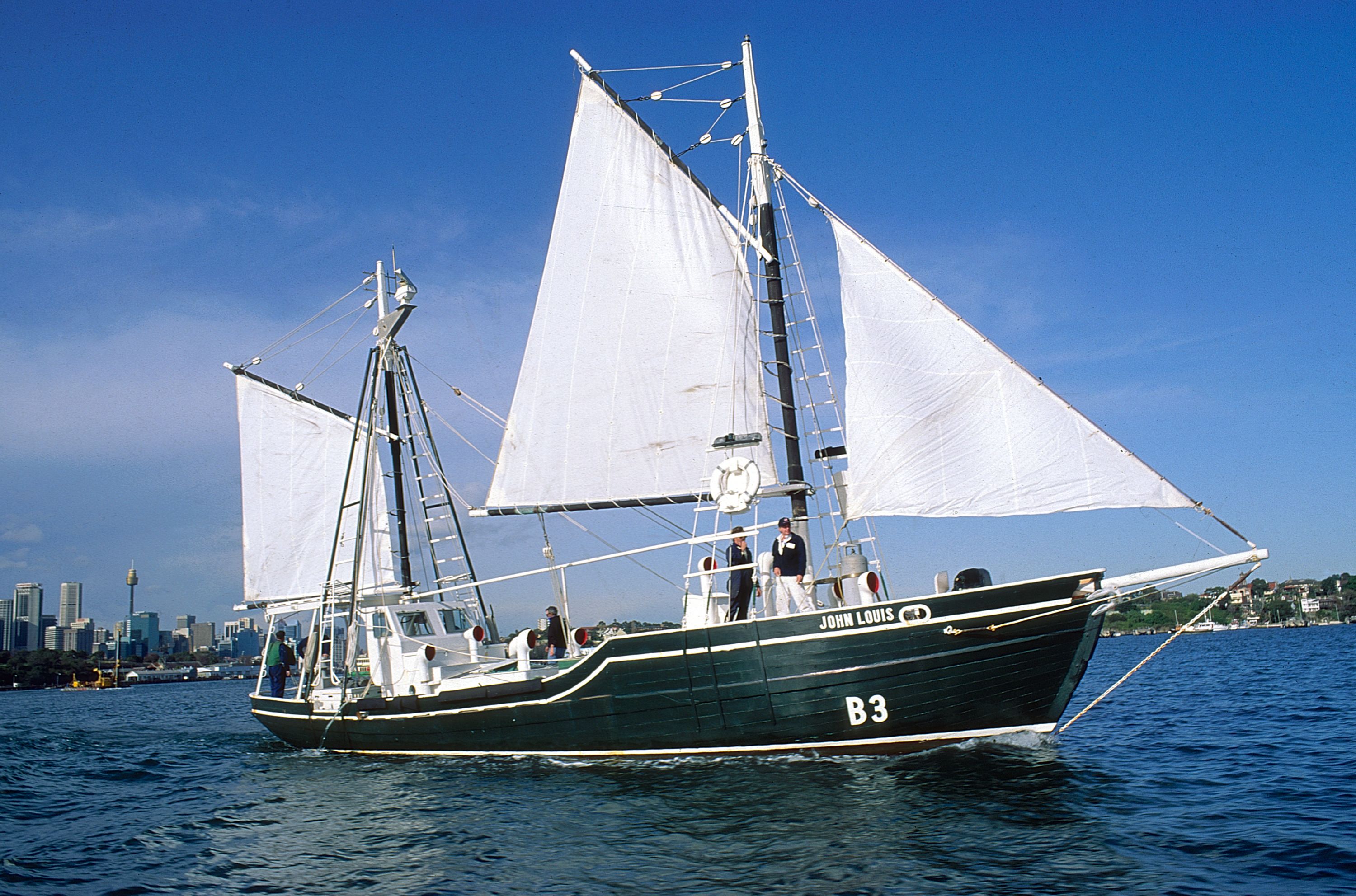 Photo of a boat with a dark green hull and white sails on blue water with a clear blue sky. 