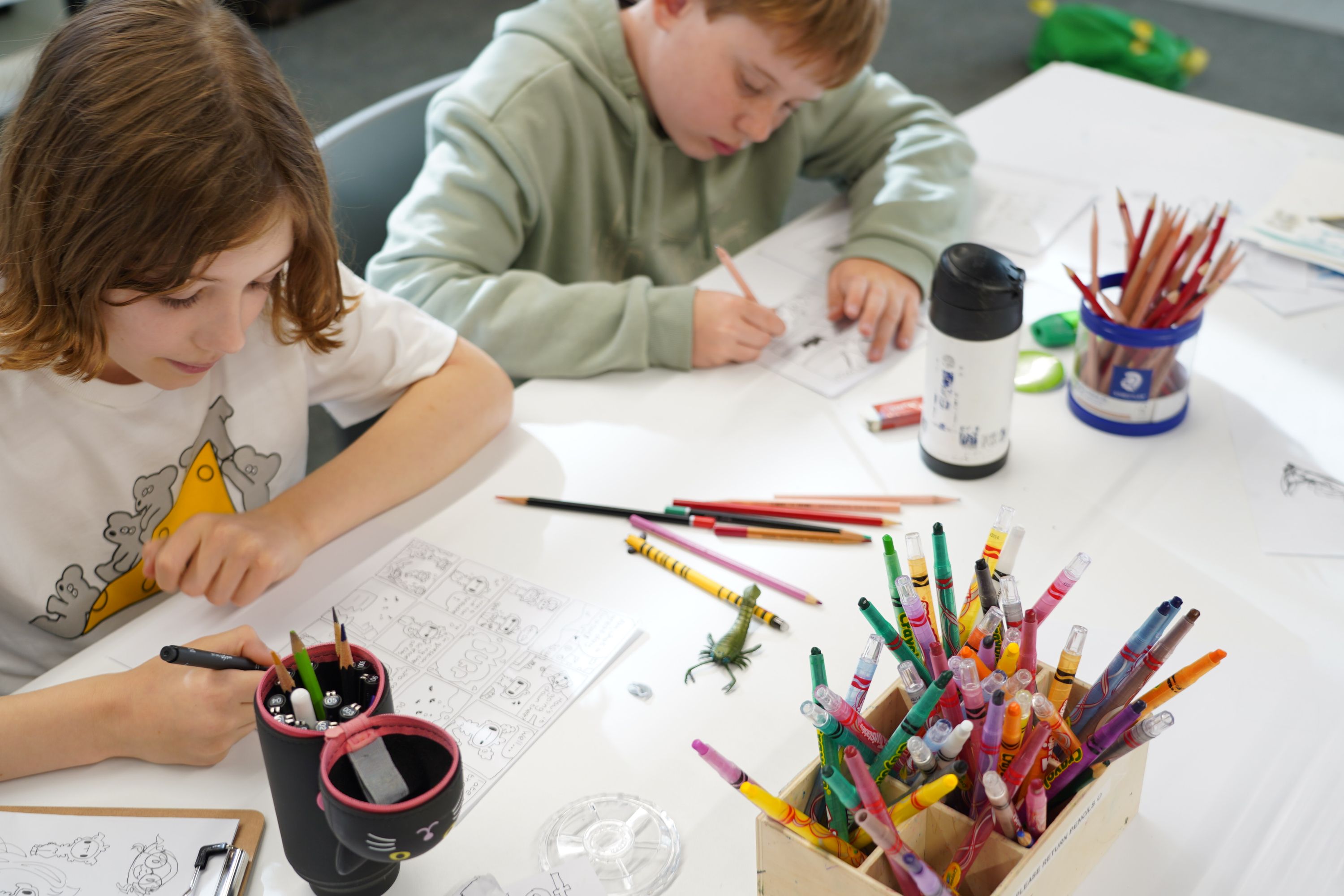 2 Children seated at a table drawing with coloured pencils.